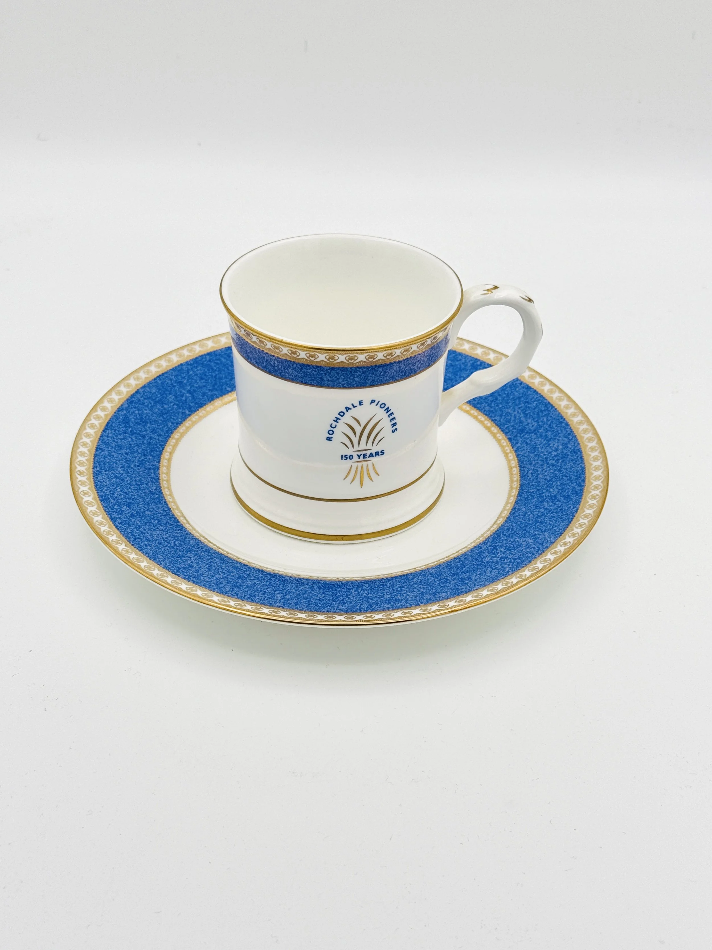 A white ceramic cup with blue and gold decorative details, featuring the logo and text 'Rochdale Pioneers ISO Years' on the front, sitting on a matching saucer with the same design, placed on a white surface.