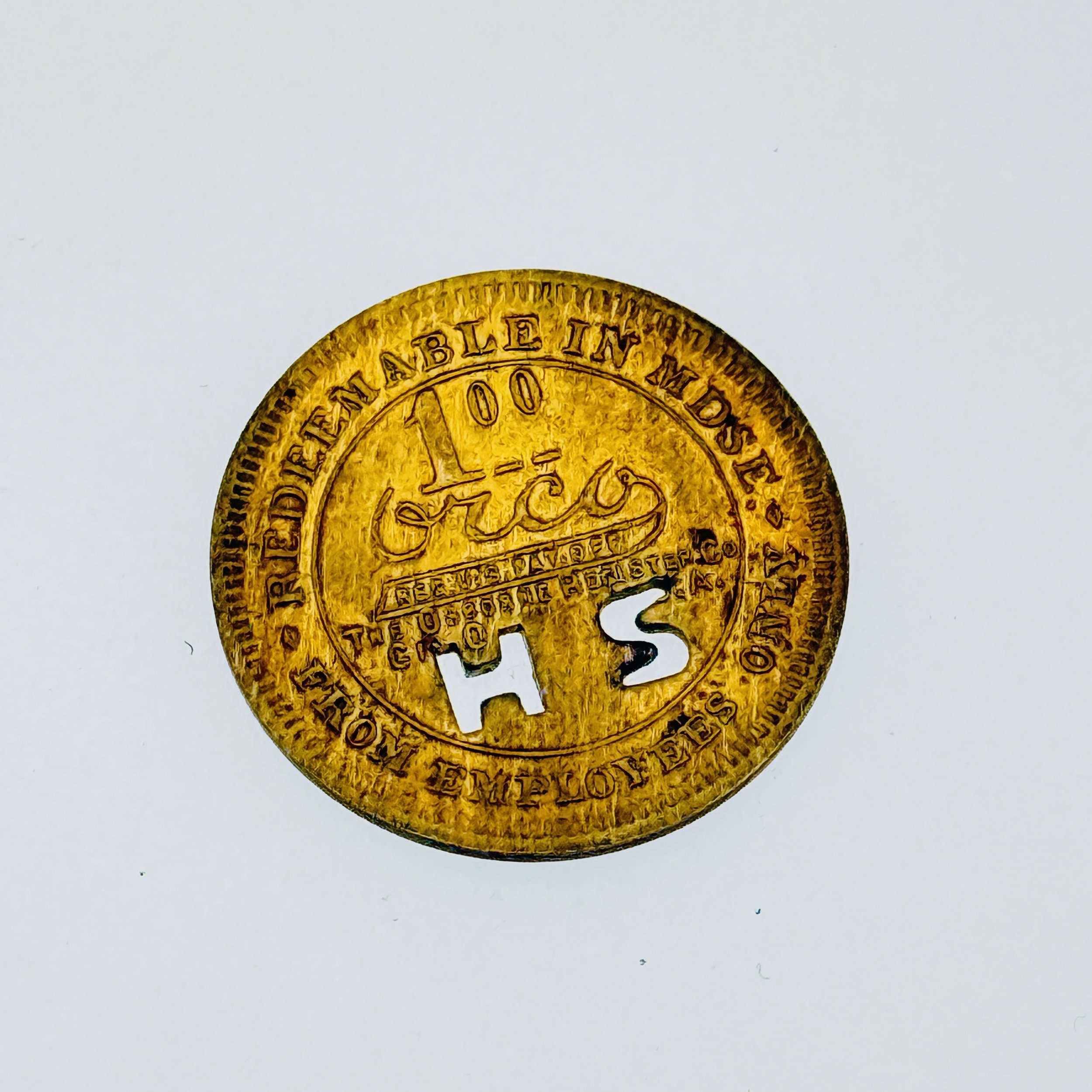Close-up of a vintage brass token with the words 'H S' cut out in the center, and inscriptions including 'REDEEMABLE IN MIDDLE' and 'THE' around the edge.