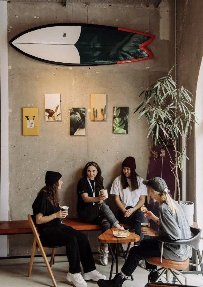 Four young women sitting and standing in a cafe, engaged in conversation, with coffee cups and a donut on a small table. The cafe has an art deco cactus display and a surfboard mounted on a textured concrete wall, with four plant-themed framed pictures.
