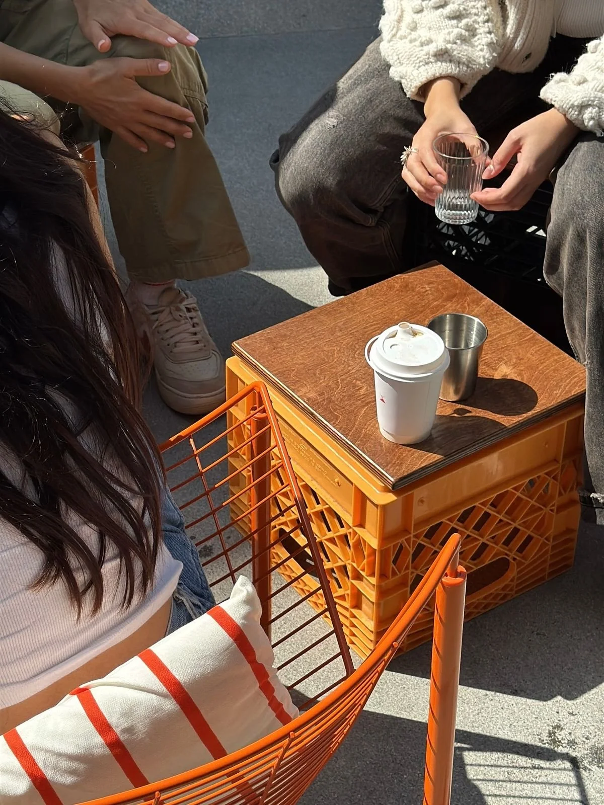 Two people sitting outdoors at a low table made of stacked orange crates. One person is holding a glass of water, another has a disposable coffee cup and a metal cup on the table. There is a pillow with orange and white stripes on an orange wire chair. The table has a wooden top and is set on a gray concrete surface.