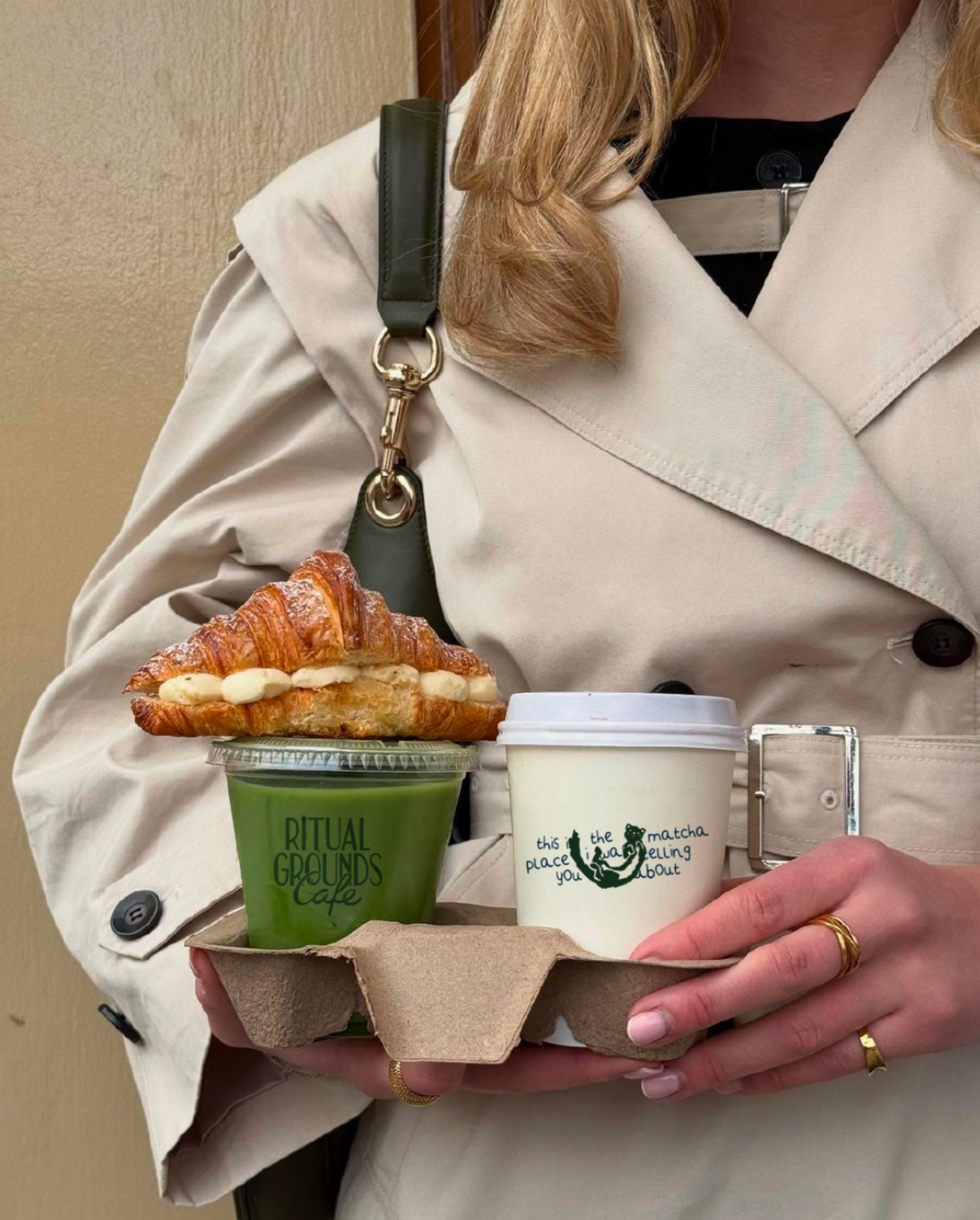 Person holding a tray with a matcha drink and a foam-topped croissant with cream filling, wearing a beige trench coat, with a brown hand, gold rings, and a black shoulder strap.