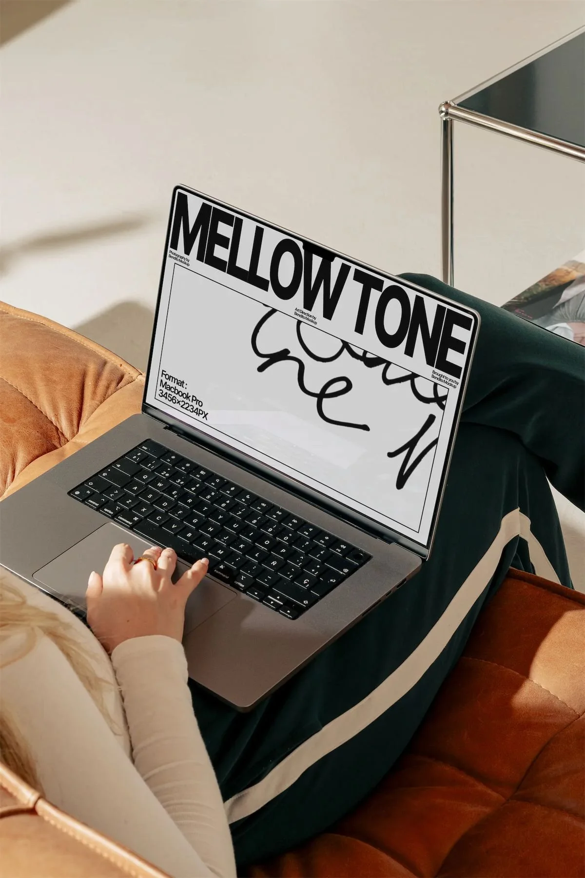 Person using a laptop with a screen displaying the word 'MELOW TONE' in large letters and a partial doodle beneath it. The person is seated on a brown couch, wearing a white long-sleeve shirt and a ring on their finger.