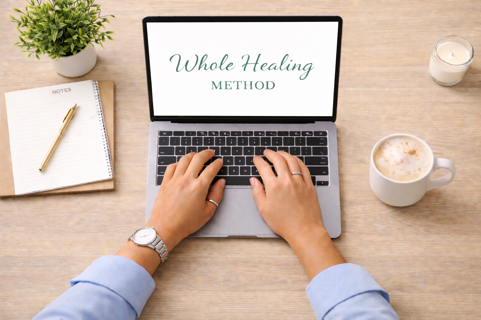 A person working on a laptop at a wooden desk with a notebook, pen, potted plant, cup of coffee, lit candle, and a mug of coffee or tea. The laptop screen displays the words 'Whole Healing Method'.