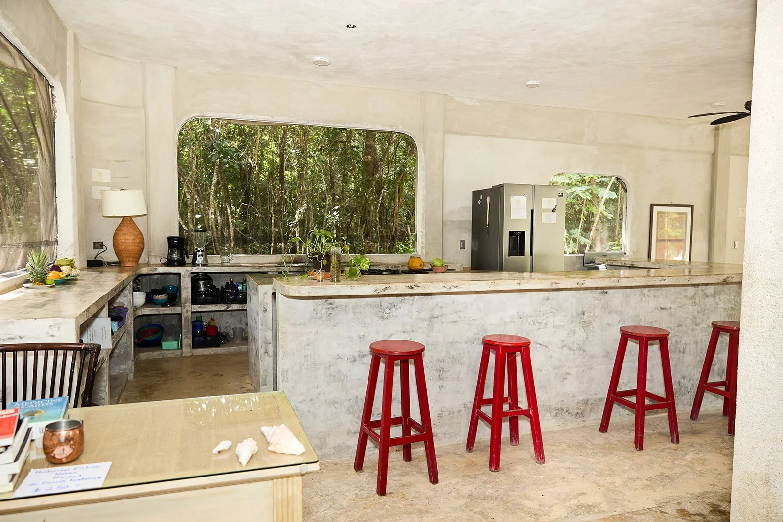 Kitchen area with white concrete walls and counter, three red bar stools at the counter, large window revealing trees outside, appliances including a double-door refrigerator, and various kitchen utensils and decorations.