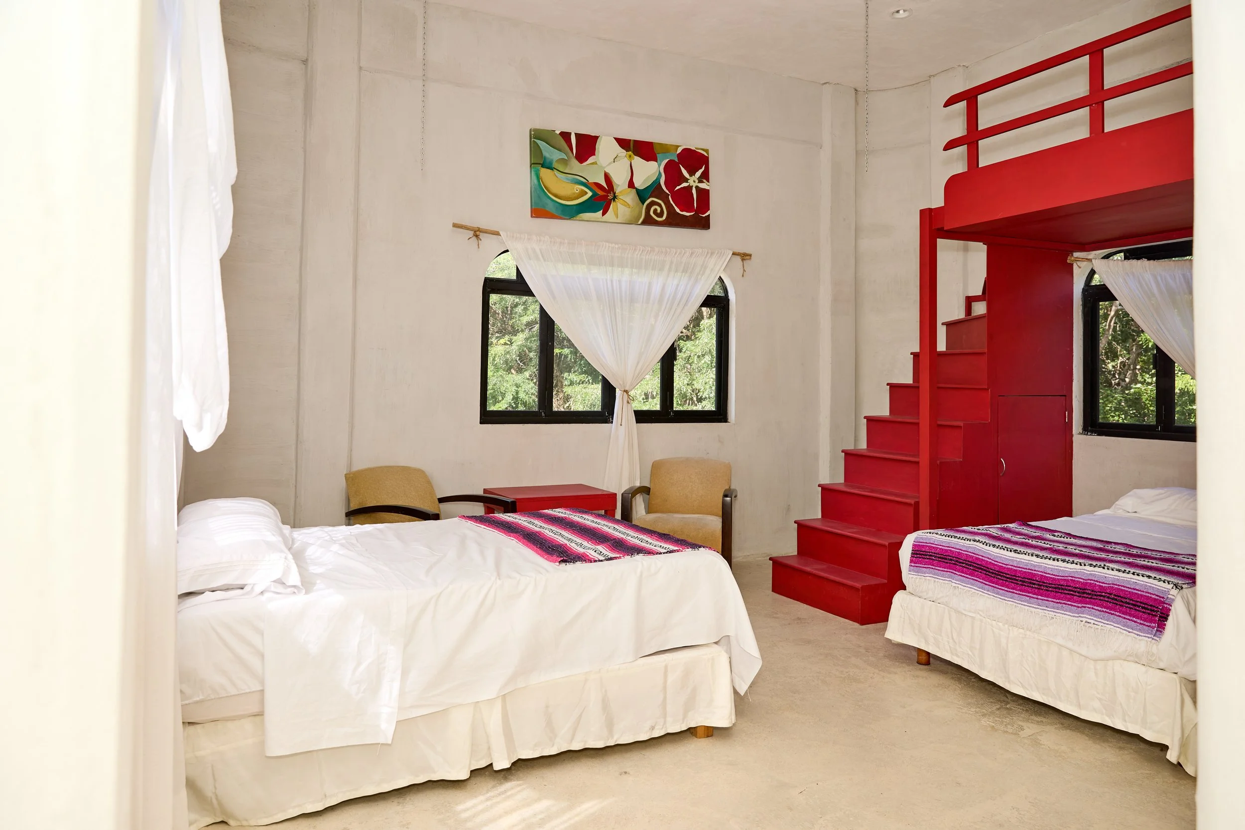 A hotel room with two beds, one with white bedding and a pink and black striped blanket, and another with white bedding and a similar blanket, separated by a red staircase leading to a loft. There are two beige chairs, a small red table, two windows 