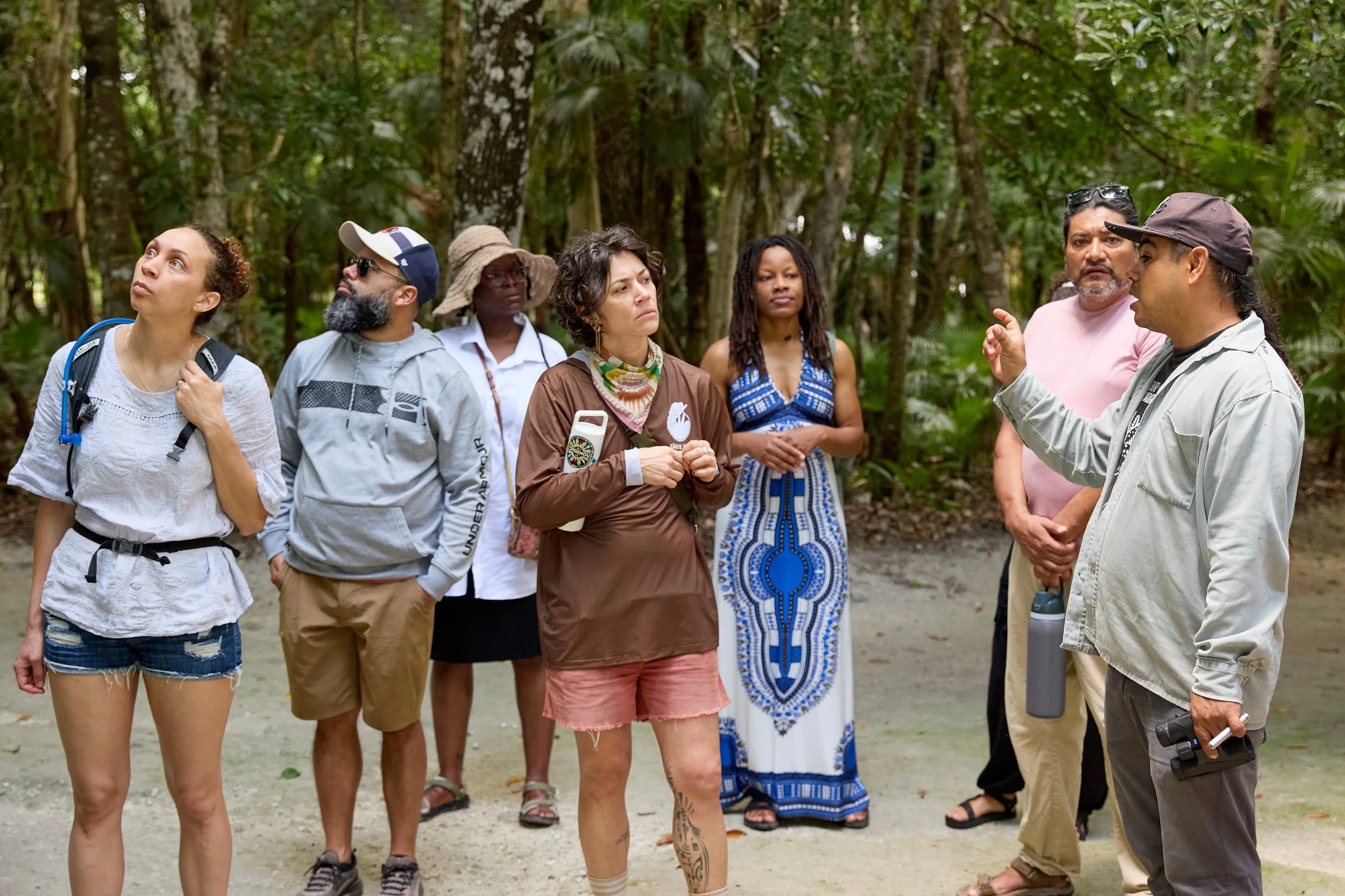 Group of people listening to a guide in a forested area, some with backpacks and hats, engaged in a discussion.