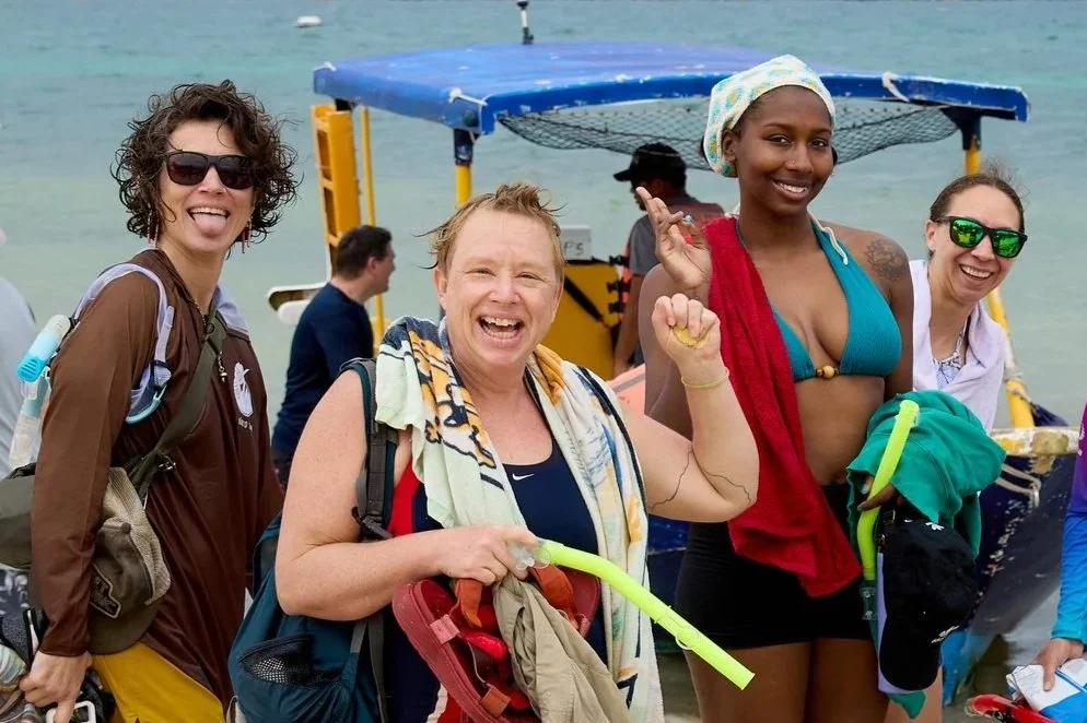 Four smiling women in swimwear and casual clothes stand together near a boat on the water. They are holding snorkeling gear, and one woman has a towel draped over her shoulder. The background shows a yellow boat with a canopy.
