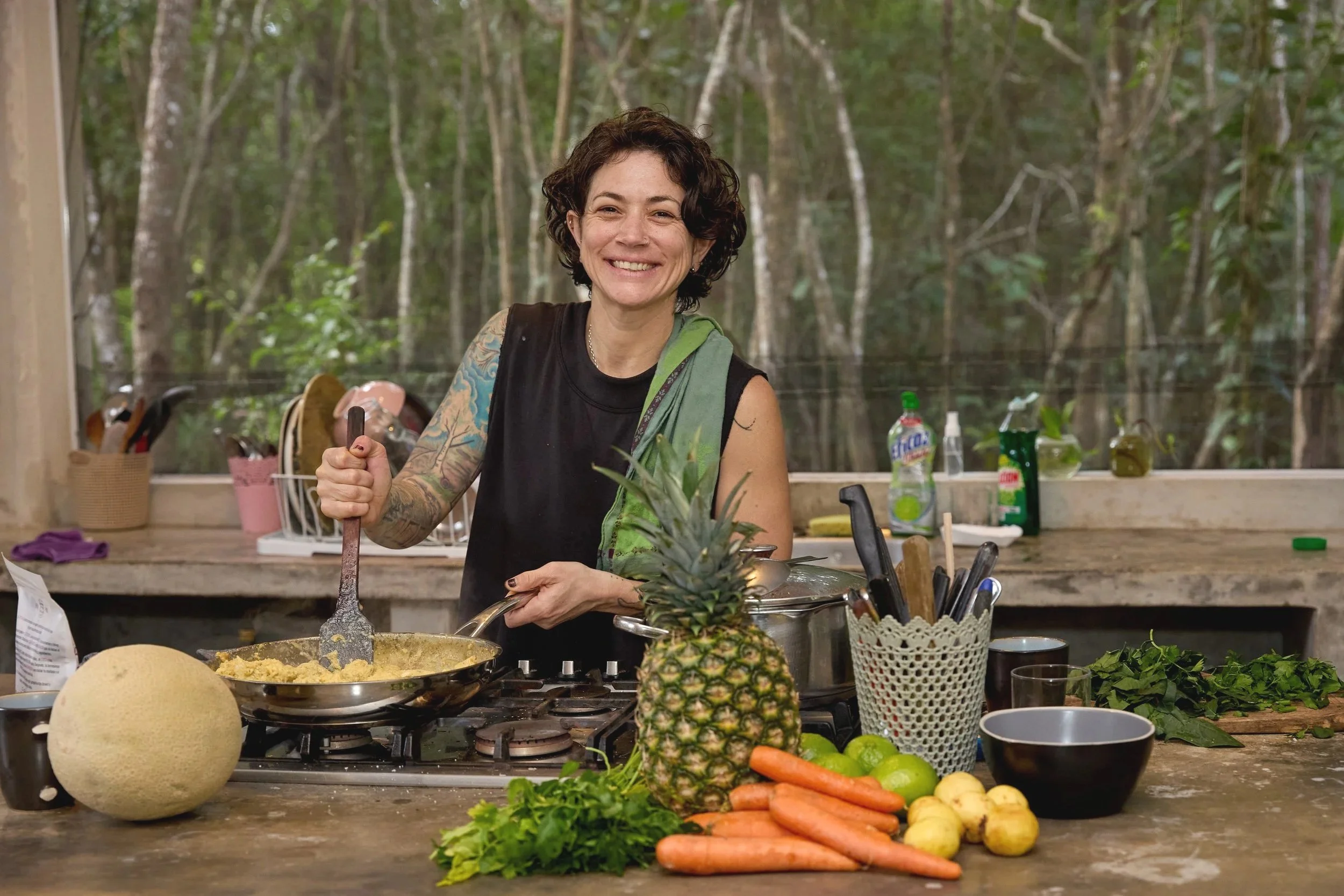 A woman with curly hair and tattoos on her arm, smiling and cooking scrambled eggs in a frying pan in an outdoor kitchen surrounded by a forest. The kitchen contains fresh vegetables, a pineapple, and various utensils and cleaning supplies.