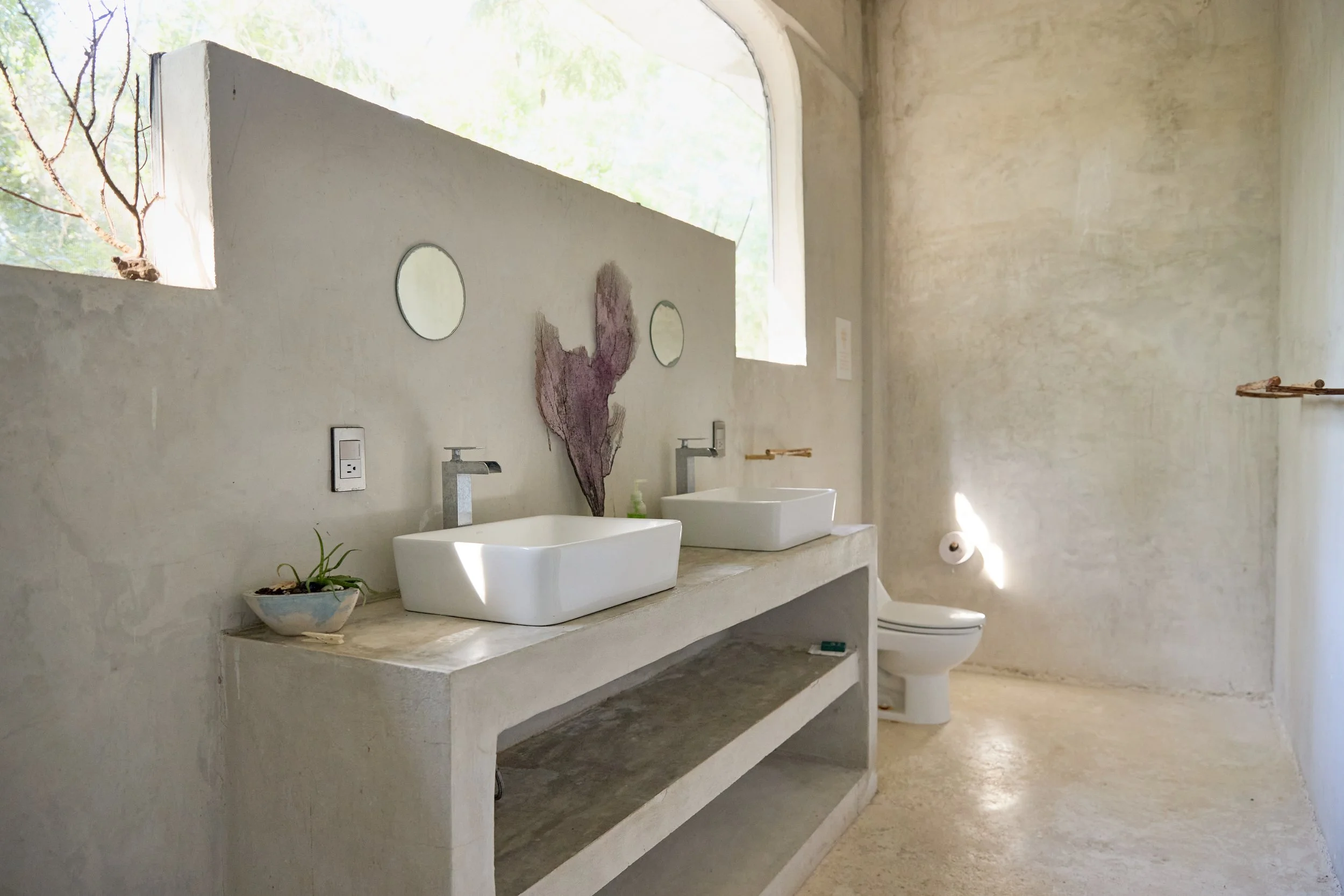 A minimalist bathroom with natural light, a double concrete vanity with two white vessel sinks, two mirrors, a small plant, and a toilet on the right side.