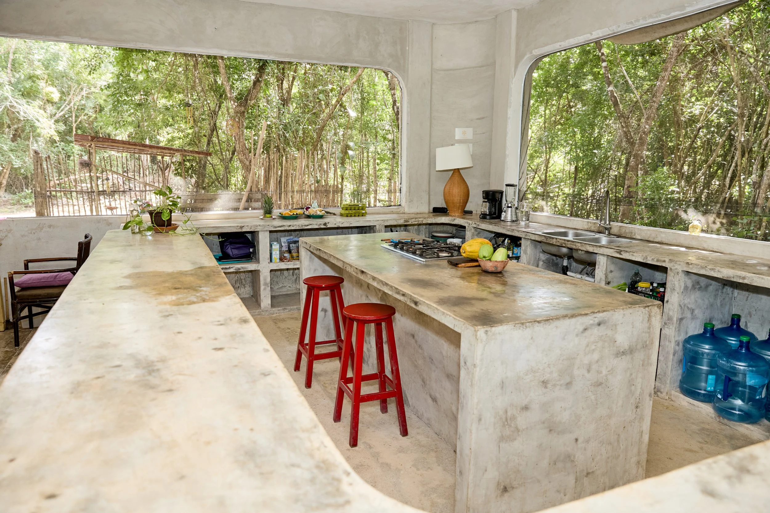 Kitchen with concrete countertops and large windows showing a forest outside, containing a stove, sink, coffee maker, water bottles, and red stools.
