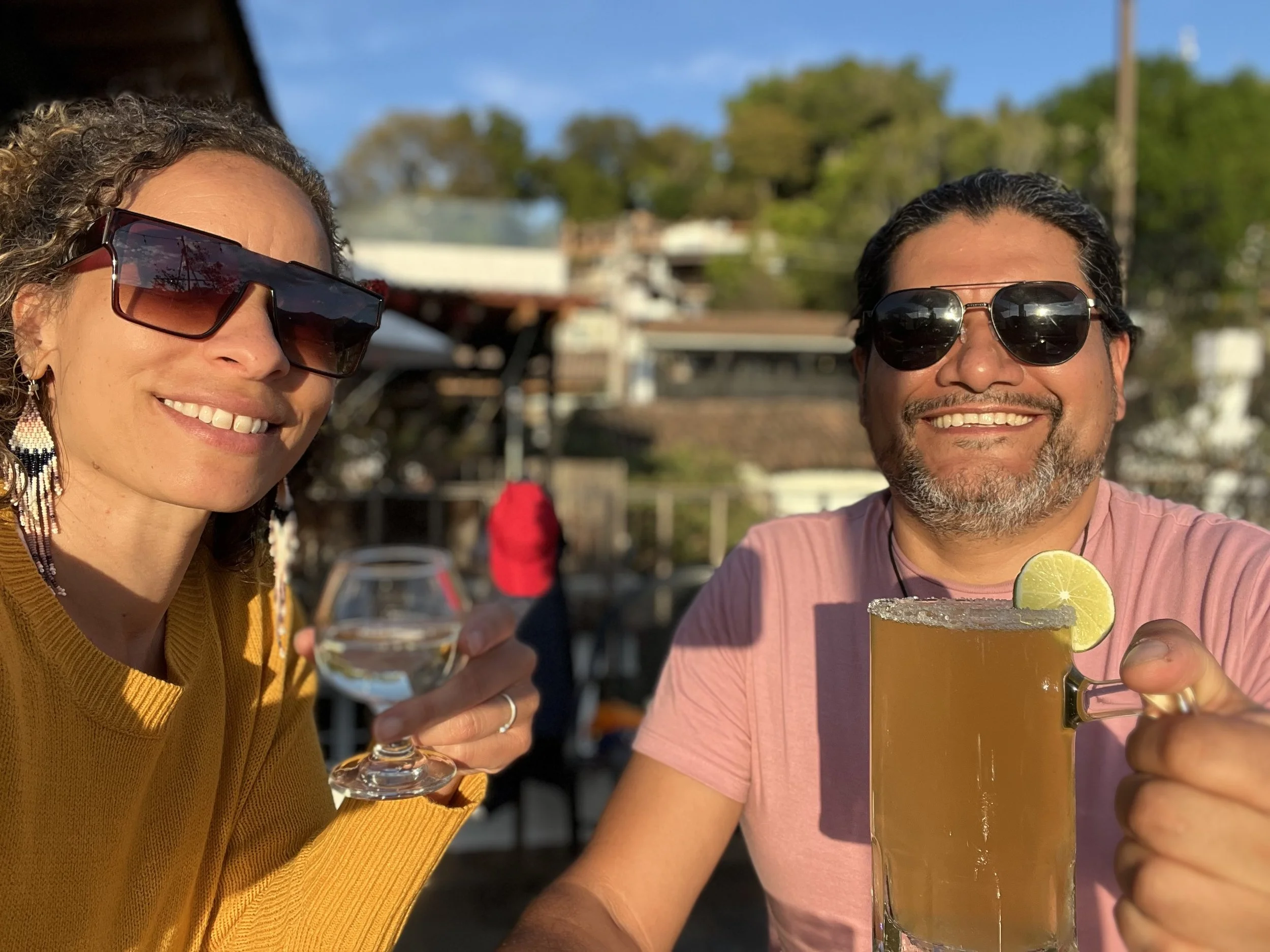 Two people smiling at a table outdoors, both wearing sunglasses. The woman on the left is holding a small glass of clear beverage, and the man on the right is holding a large glass mug with a yellow drink and a lime slice on the rim.