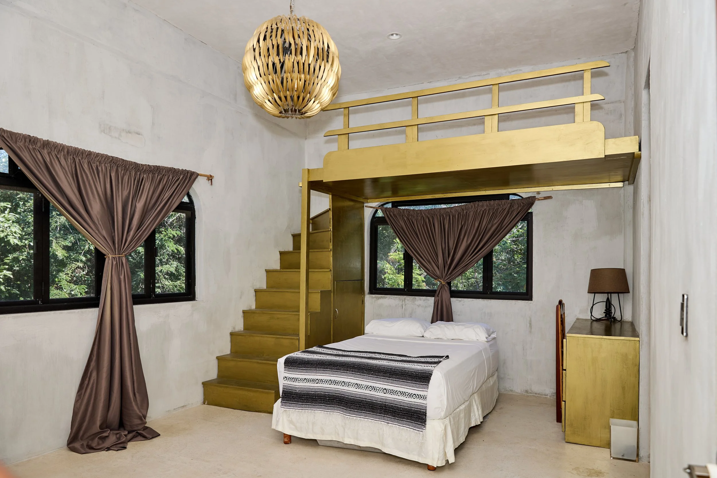 Bedroom with white walls, large windows with brown curtains, a bed with white sheets and a striped black and white blanket, a small yellow nightstand with a brown lamp, a wooden staircase leading to a lofted area, and a decorative ceiling light.