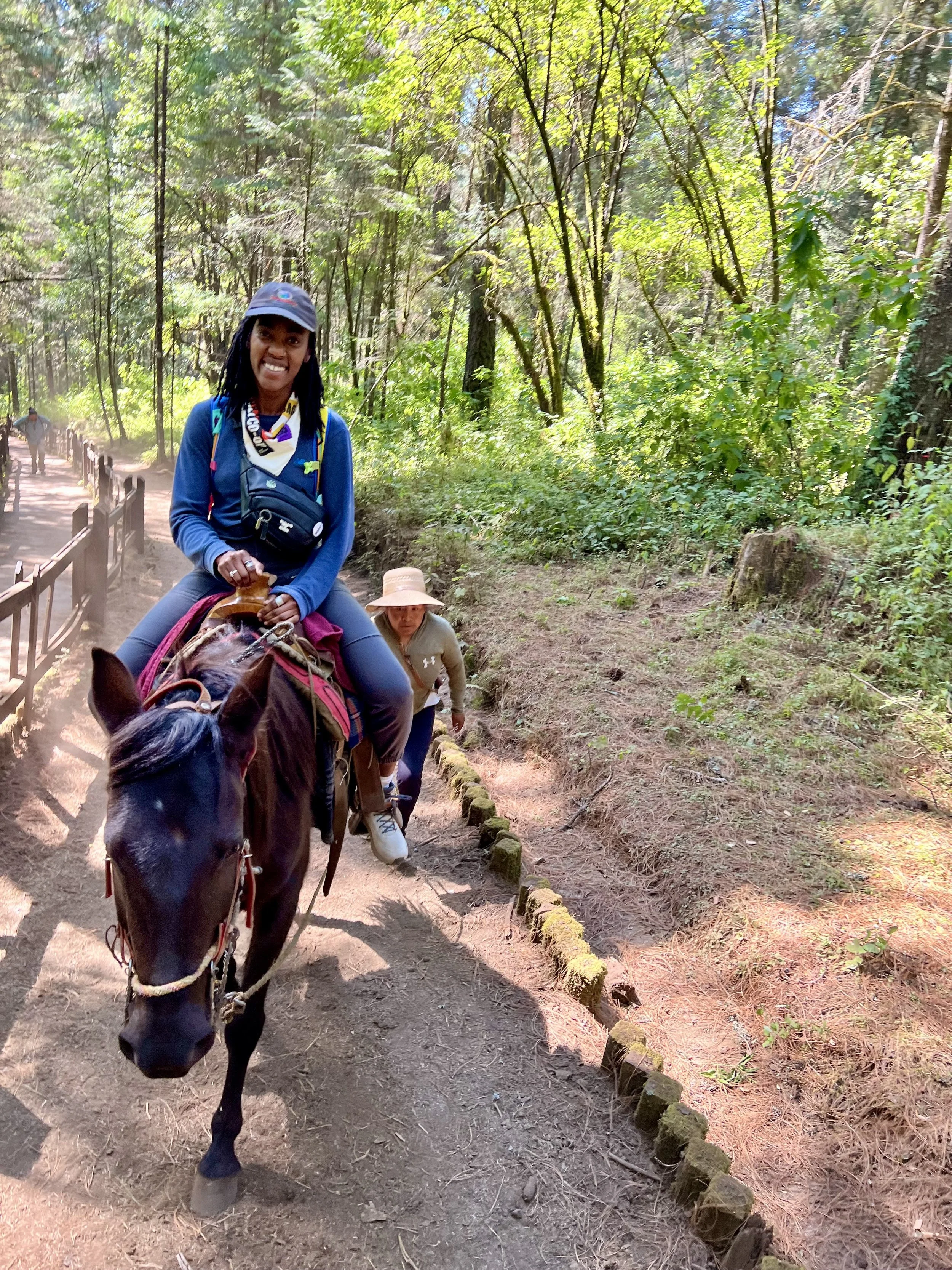 A woman riding a horse on a forest trail, with another woman walking beside her. The forest features tall trees and dense green foliage.
