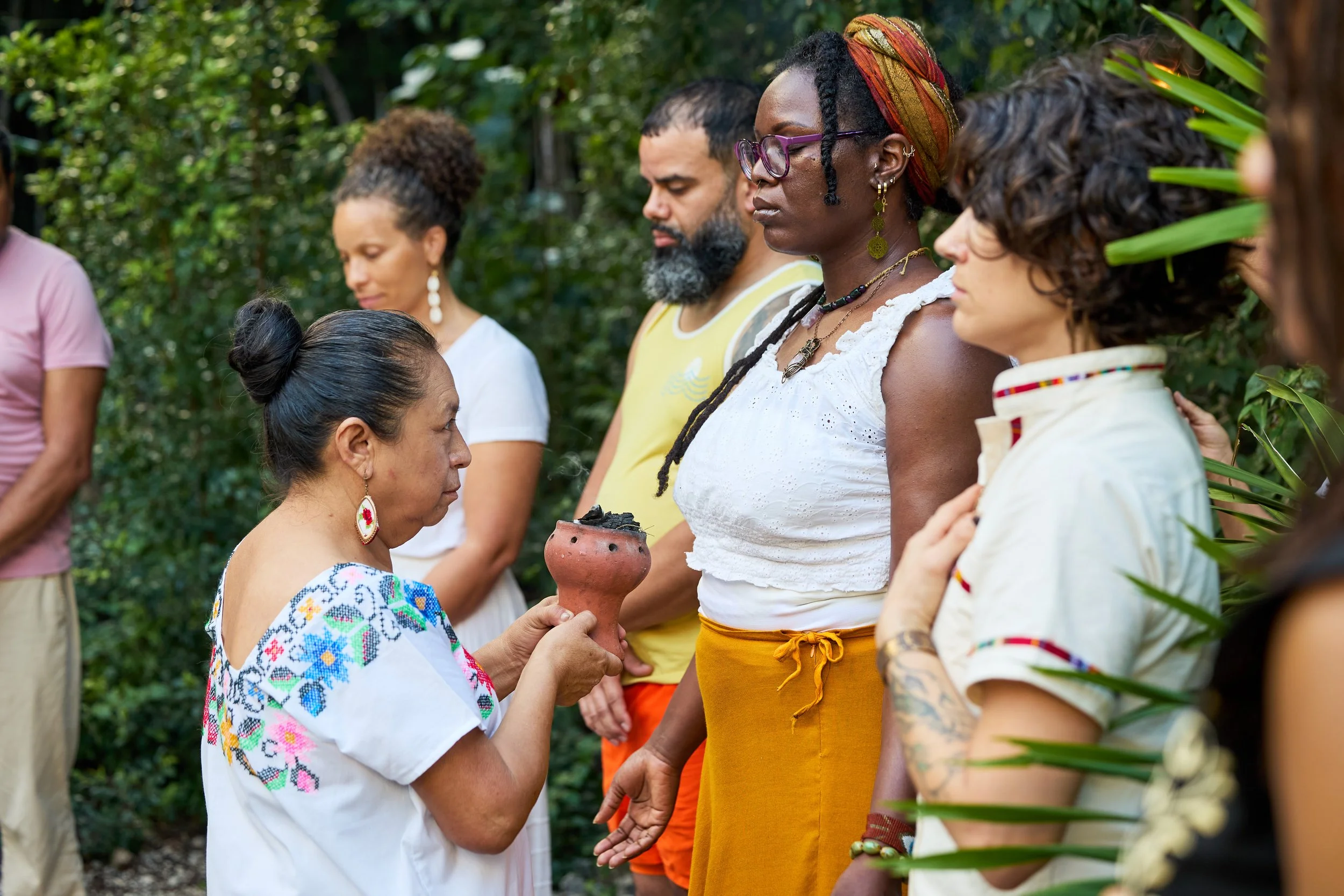 Group of diverse people participating in a spiritual or ceremonial ritual outdoors, with a woman holding a clay vessel with ashes or charcoal, others standing solemnly with eyes closed or focused, surrounded by green foliage.
