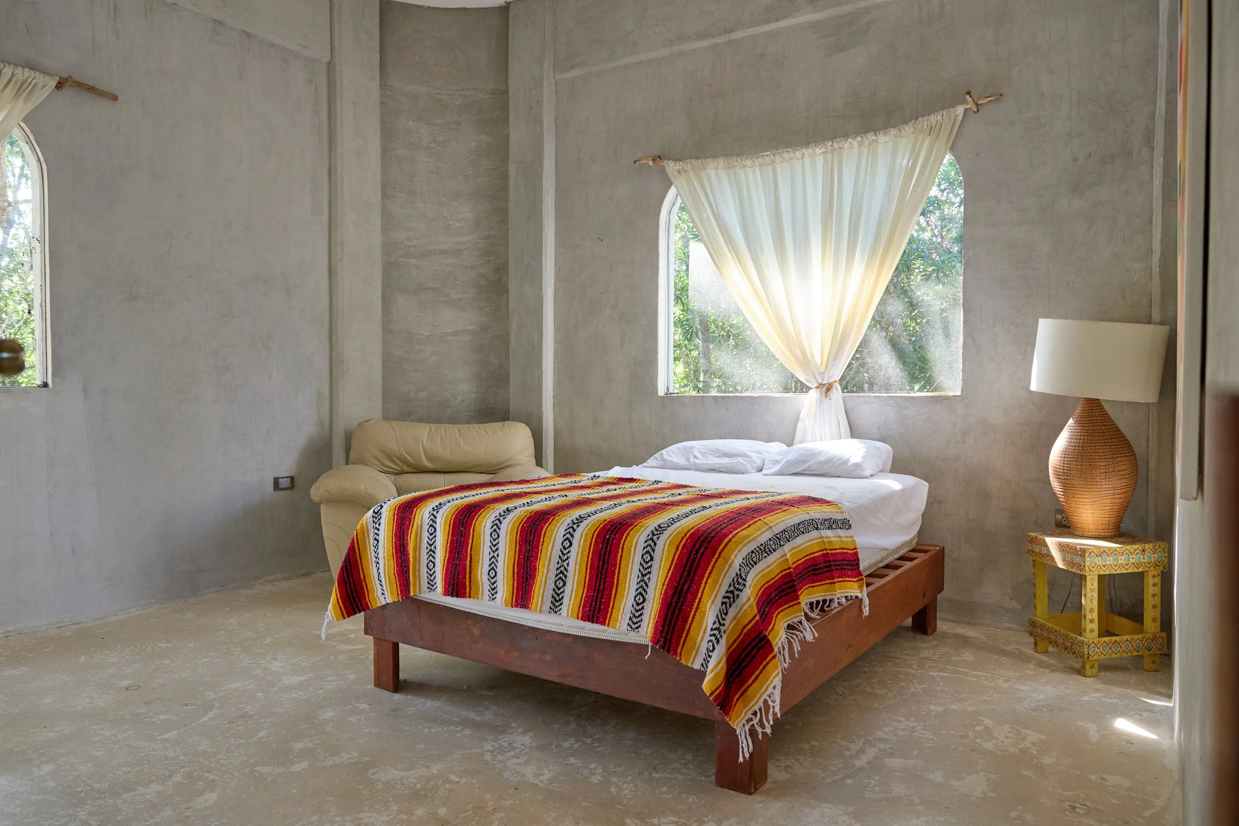 A simple bedroom with a bed covered in a colorful striped blanket, beige armchair, and a yellow side table with a large wicker lamp, light curtains, and natural sunlight streaming through window.
