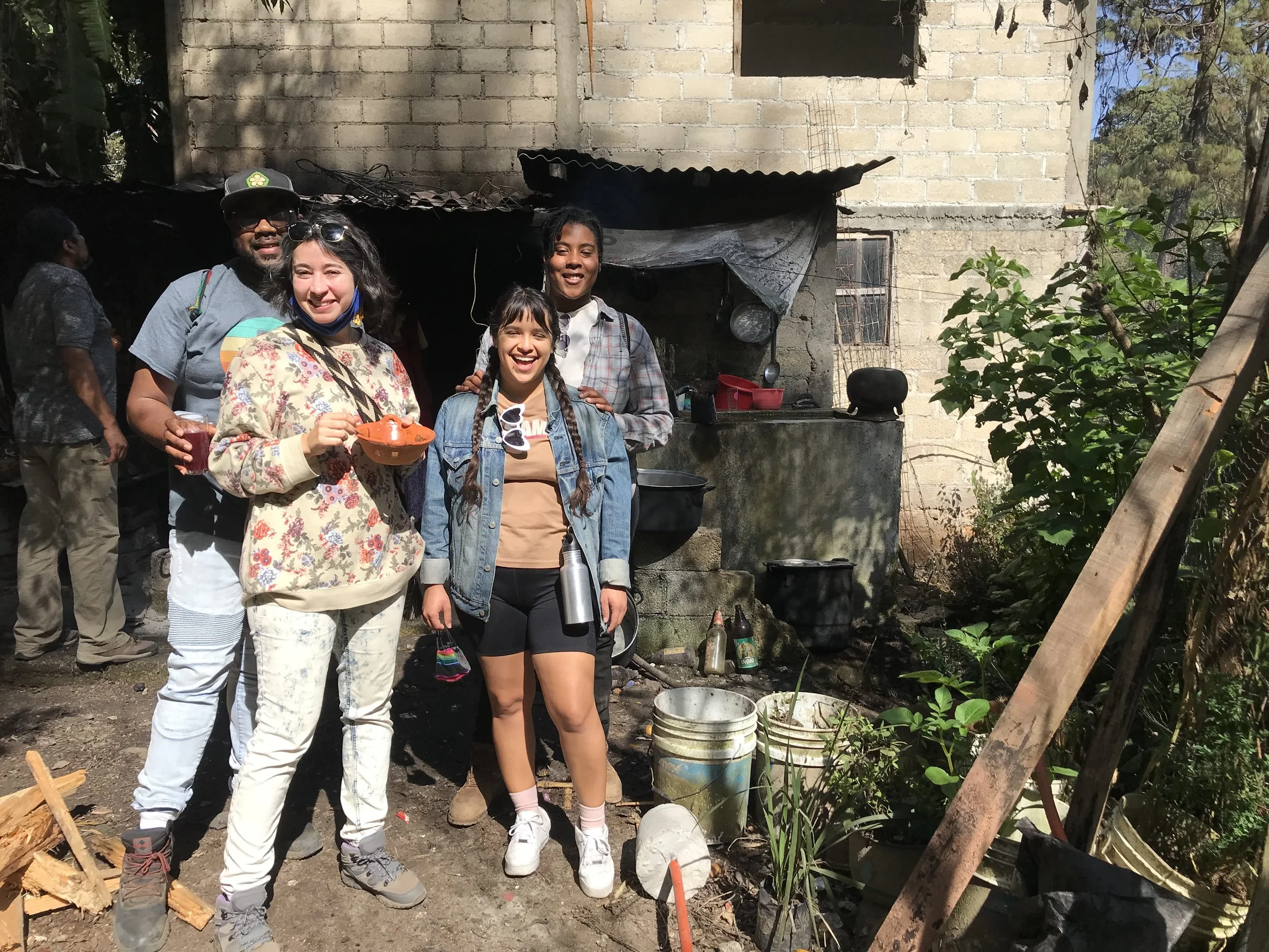 Group of four smiling young people standing outside near a rustic food stand or outdoor kitchen with an unfinished brick building in the background.