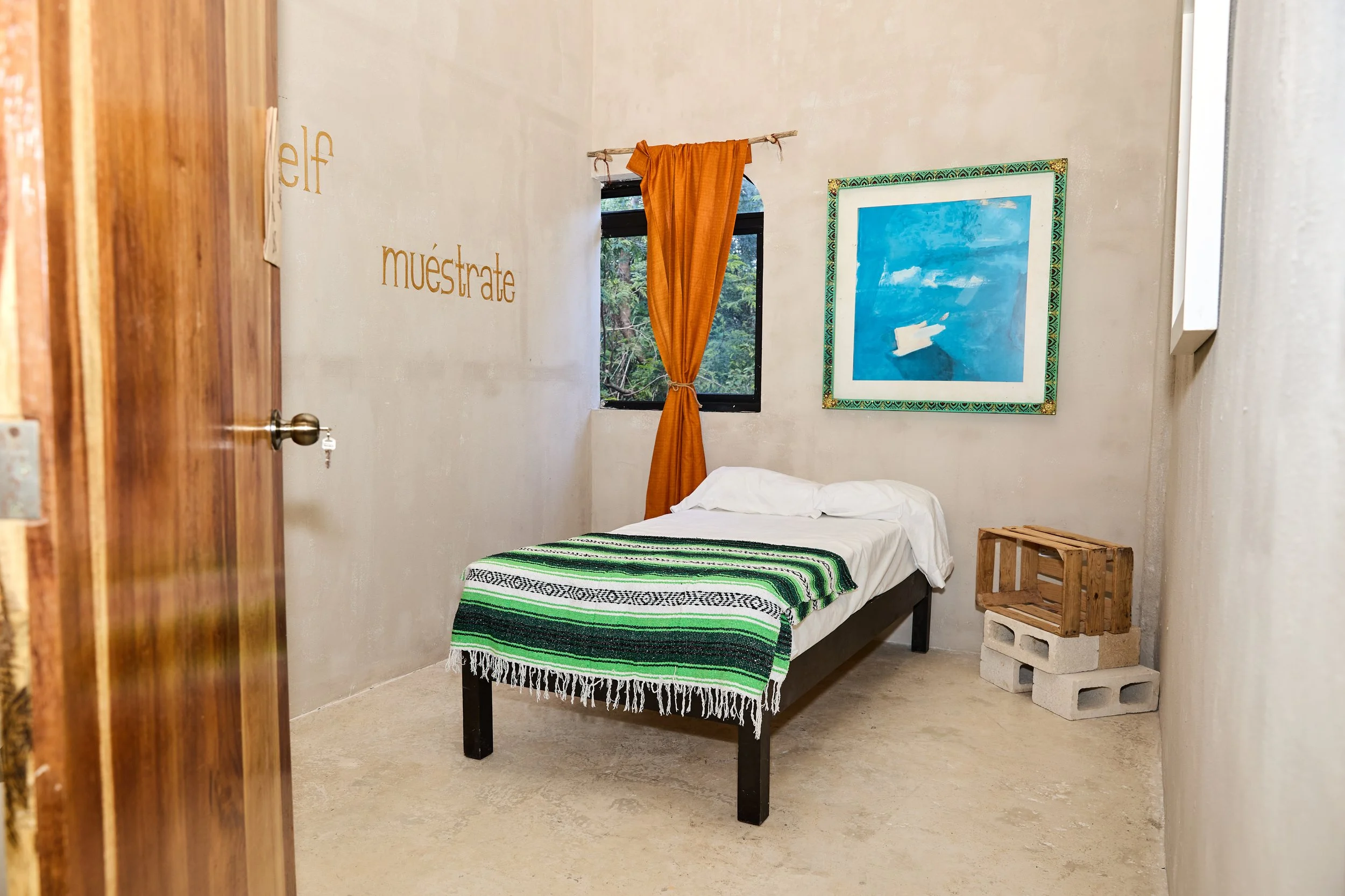 Minimalist bedroom featuring a single bed with a green, black, and white striped blanket, a small window with an orange curtain, a framed blue and white abstract painting, and a small wooden crate on concrete blocks in a beige-walled room.
