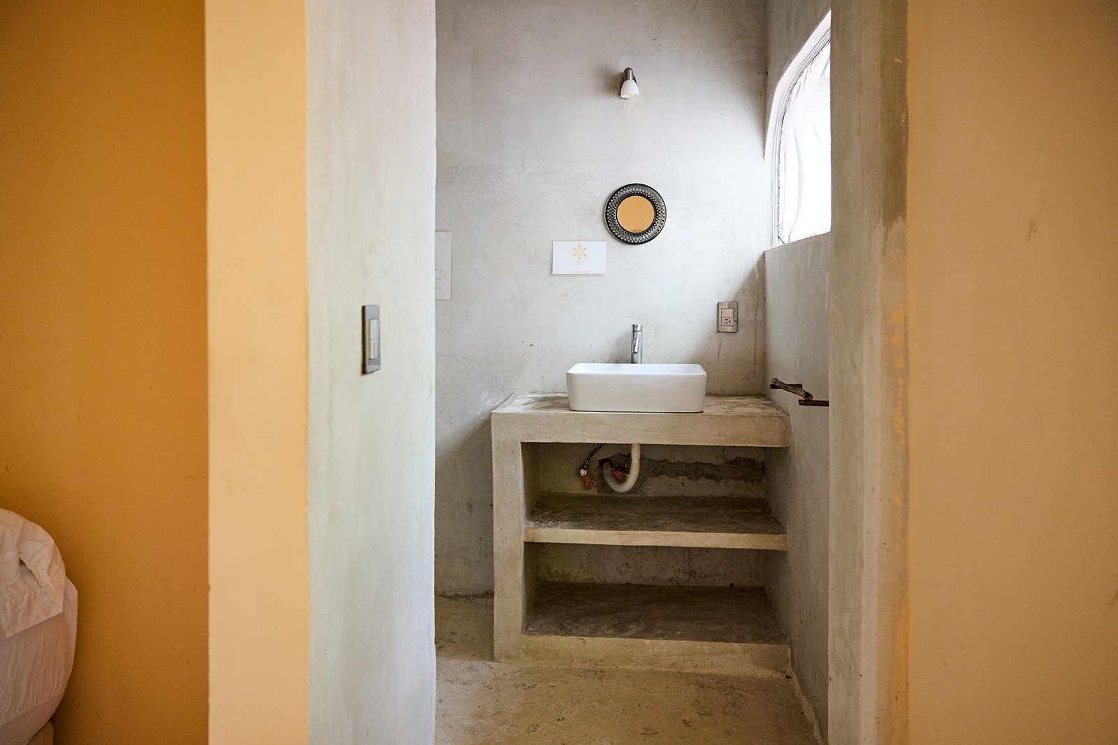 A minimalist bathroom with a concrete sink, exposed plumbing, a small shelf underneath, and a round mirror on the wall, illuminated by natural light from a nearby window.