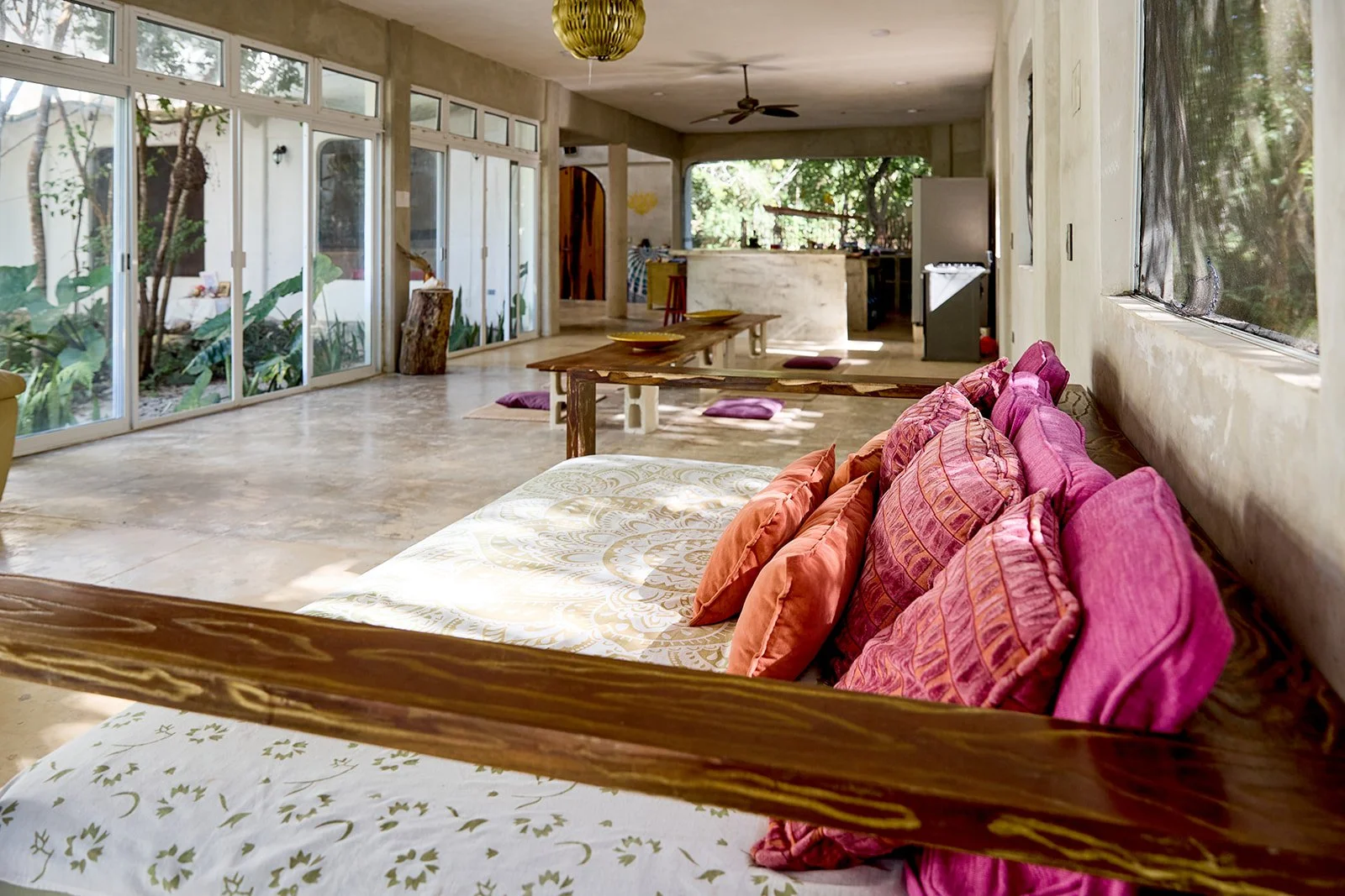 Spacious, airy living room with large glass sliding doors revealing lush greenery outside. Interior features a colorful sofa with pink and orange cushions, a low wooden table, and a concrete kitchen area in the background.