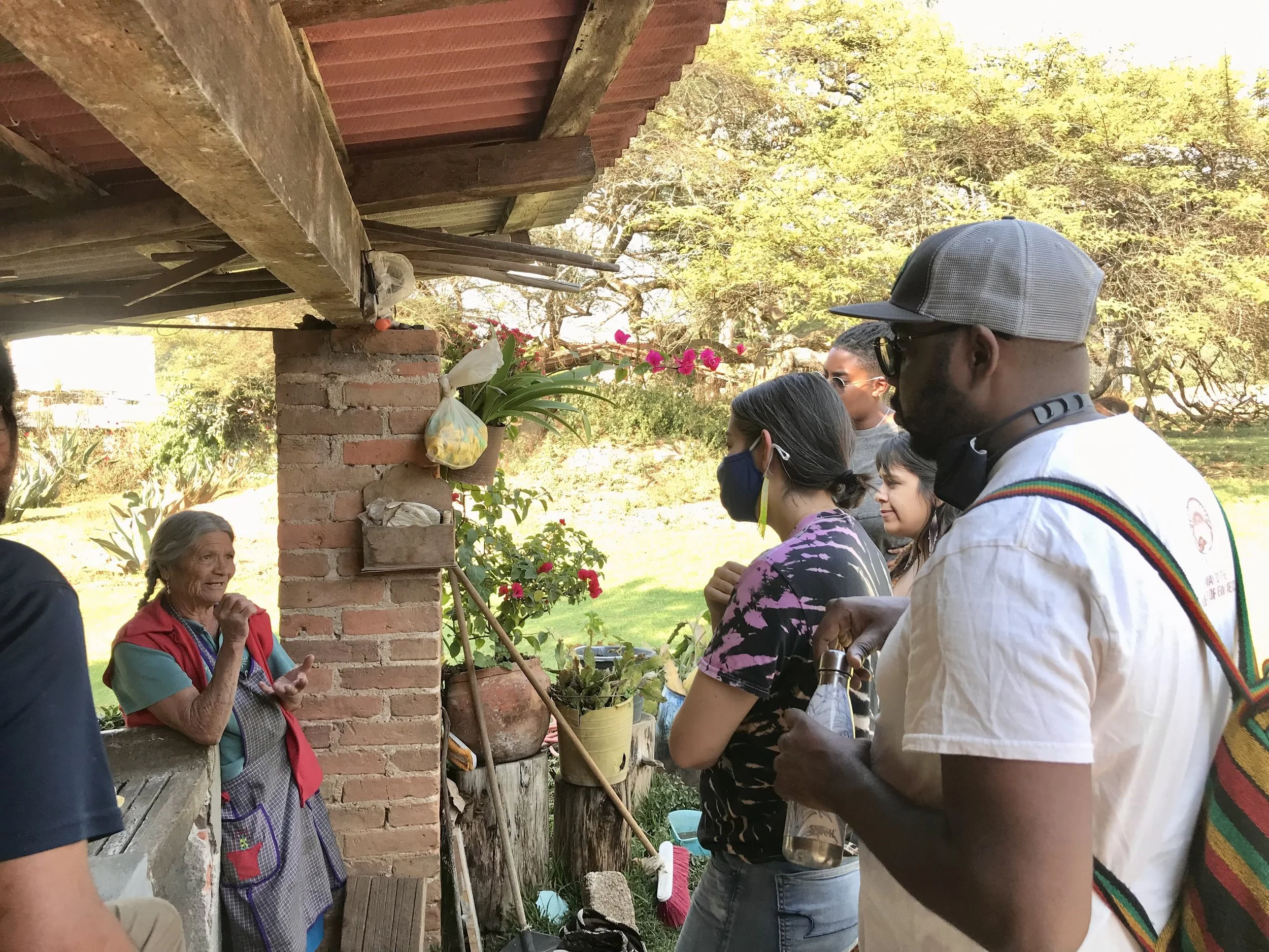 An elderly woman is speaking to a group of young adults outside near a brick building with plants and trees in the background.
