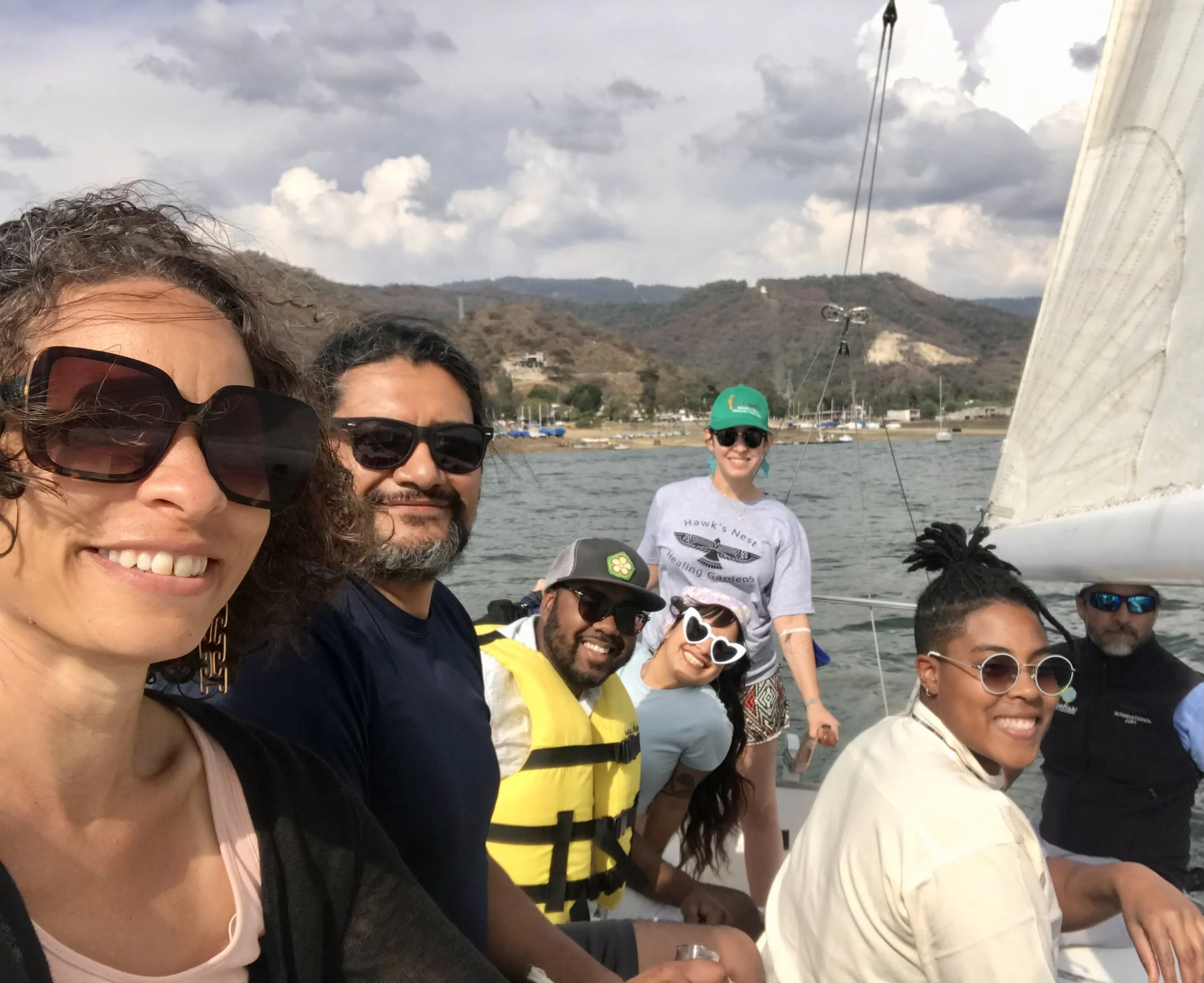 Group of six people smiling on a boat, with mountains and boats in the background, under partly cloudy sky.