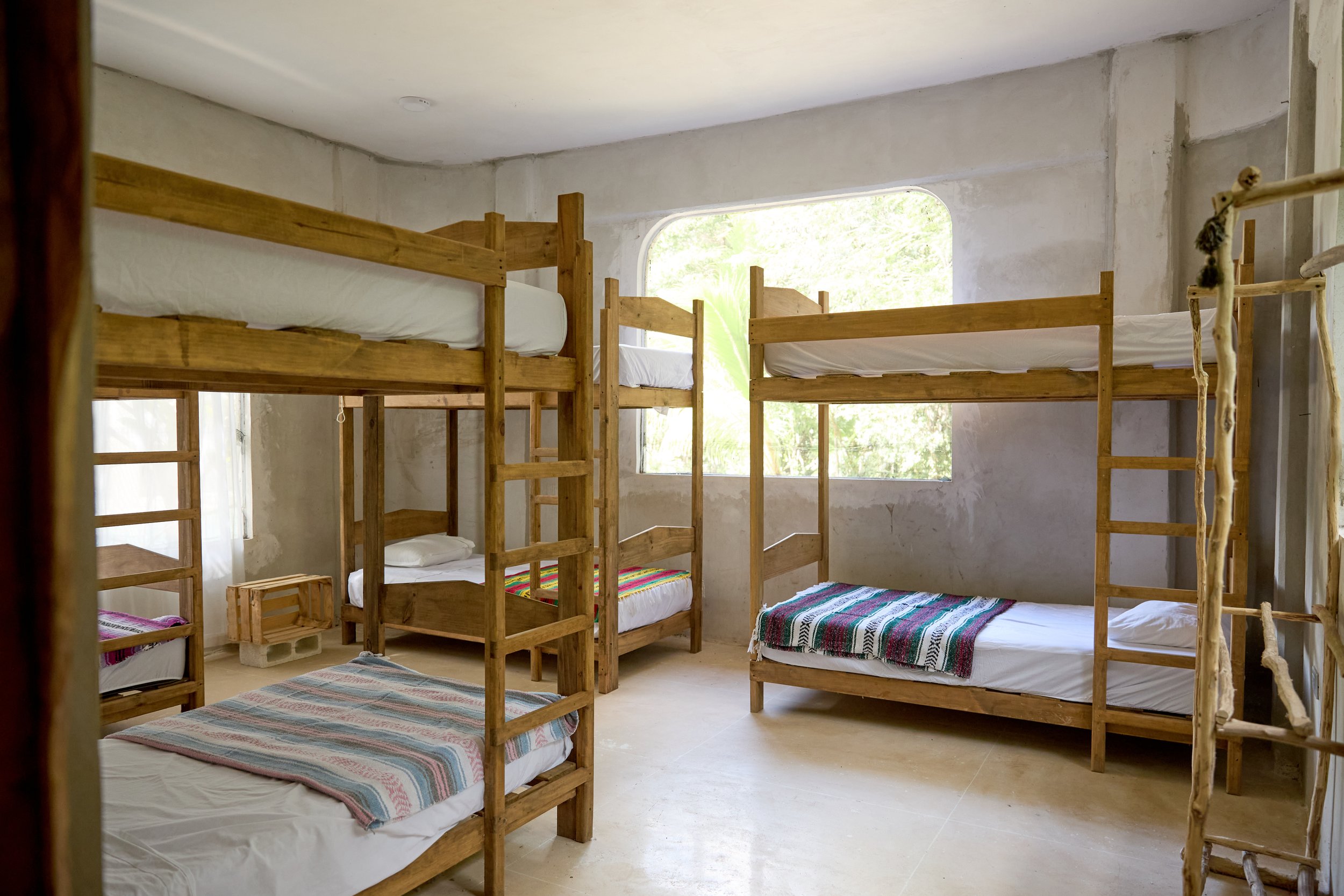 Room with multiple wooden bunk beds, some with colorful blankets, and a large window showing greenery outside.