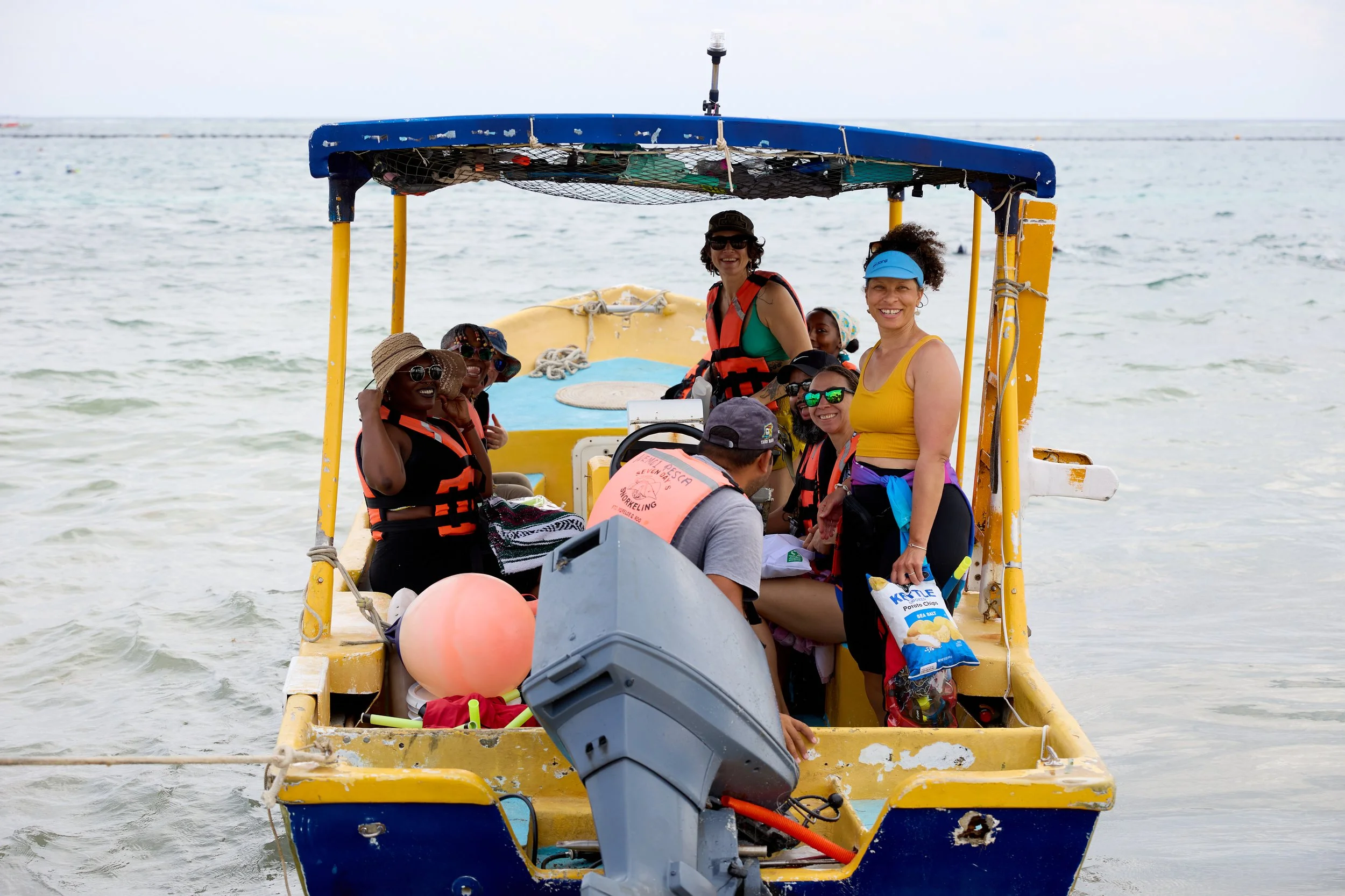 A group of people on a yellow boat in the water, wearing life jackets and summer clothing, smiling and enjoying a day at the beach.