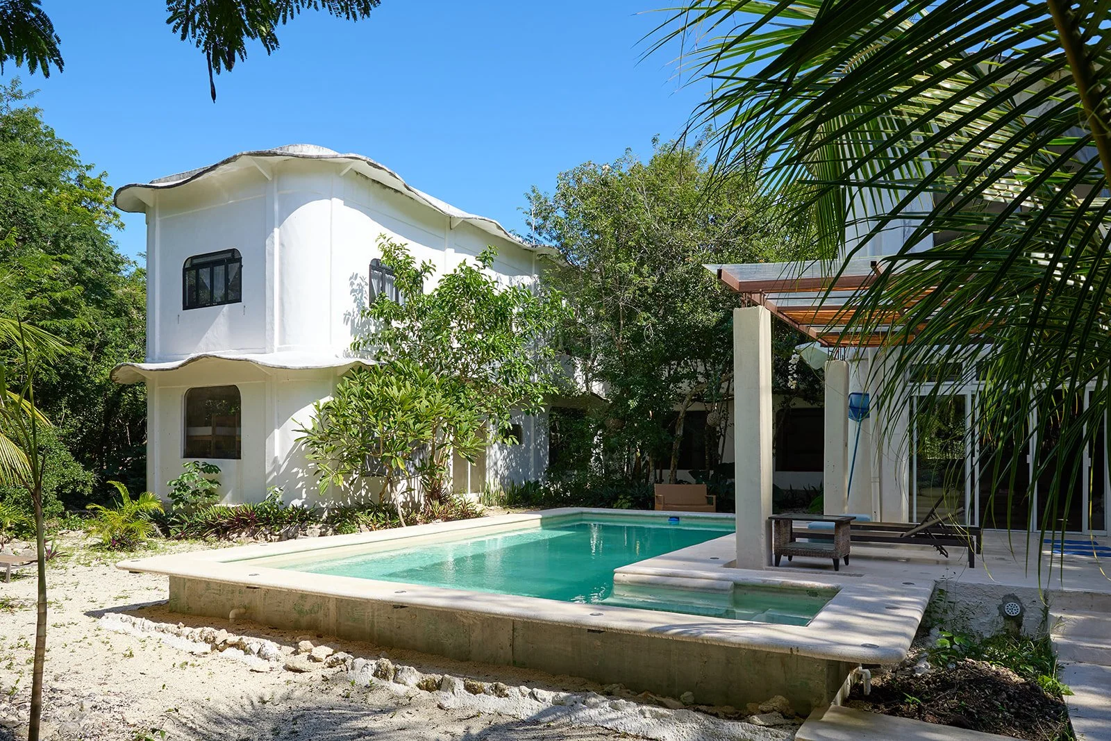 A modern two-story white house with curved architectural design, surrounded by green trees and tropical plants, with a small swimming pool in the backyard under a clear blue sky.