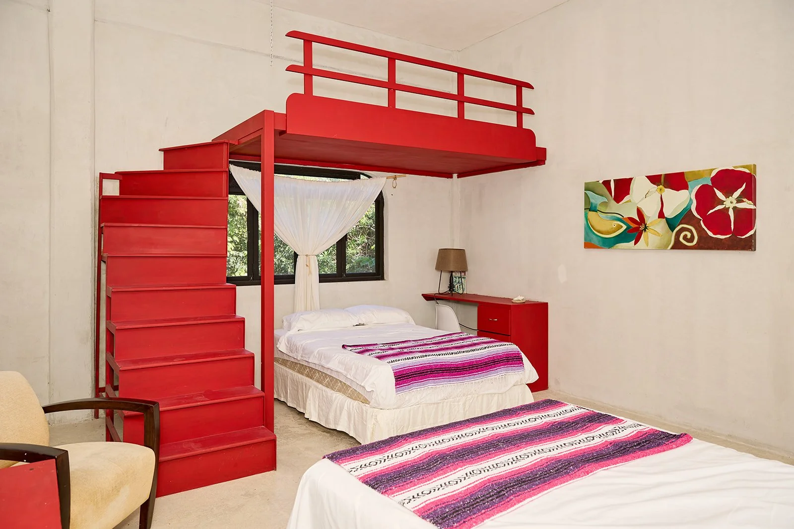 Room with two beds, a red staircase leading to a loft, a window with white curtains, colorful wall art, and beige walls.