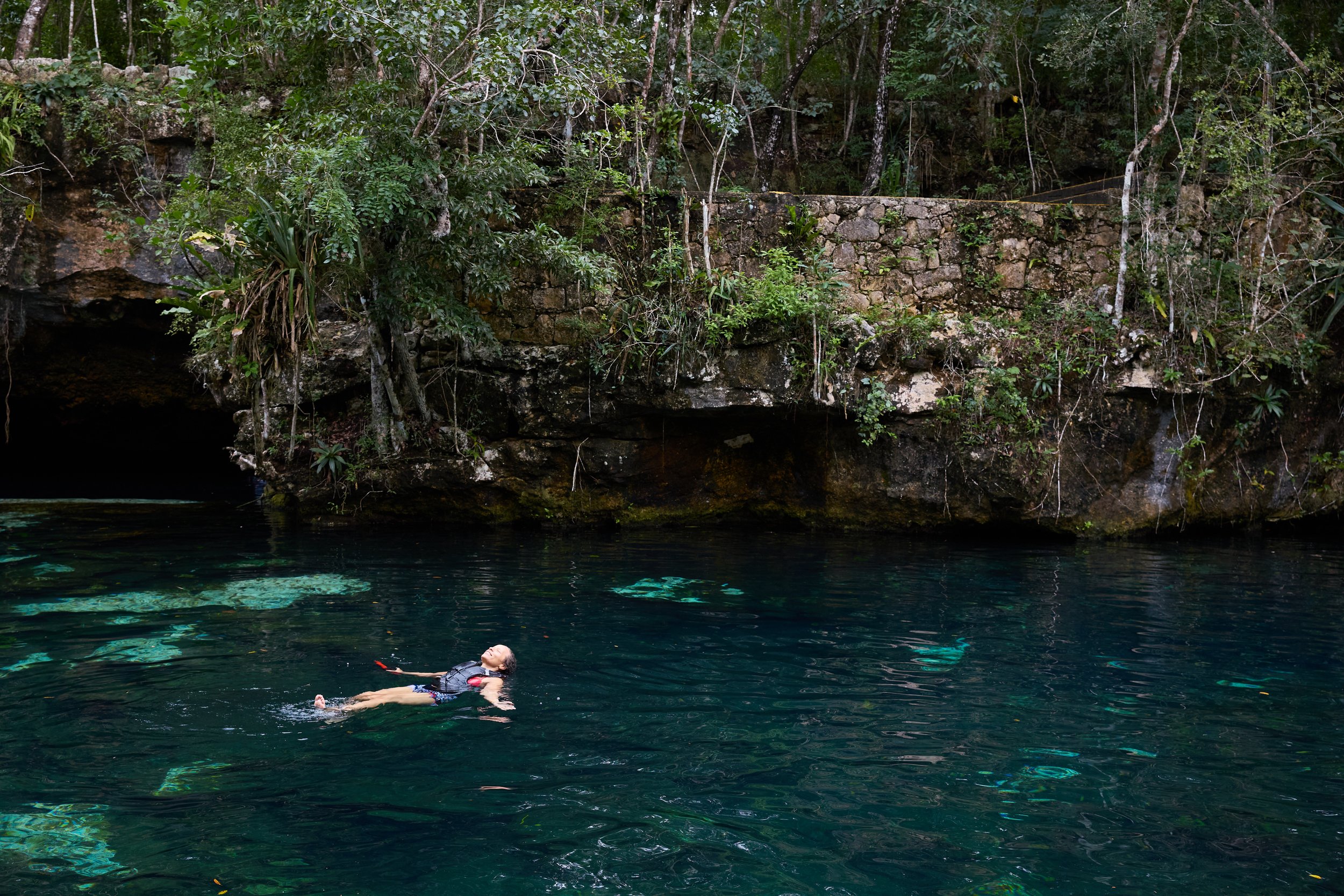 A person floating on water near a rocky and wooded shoreline in a natural setting.