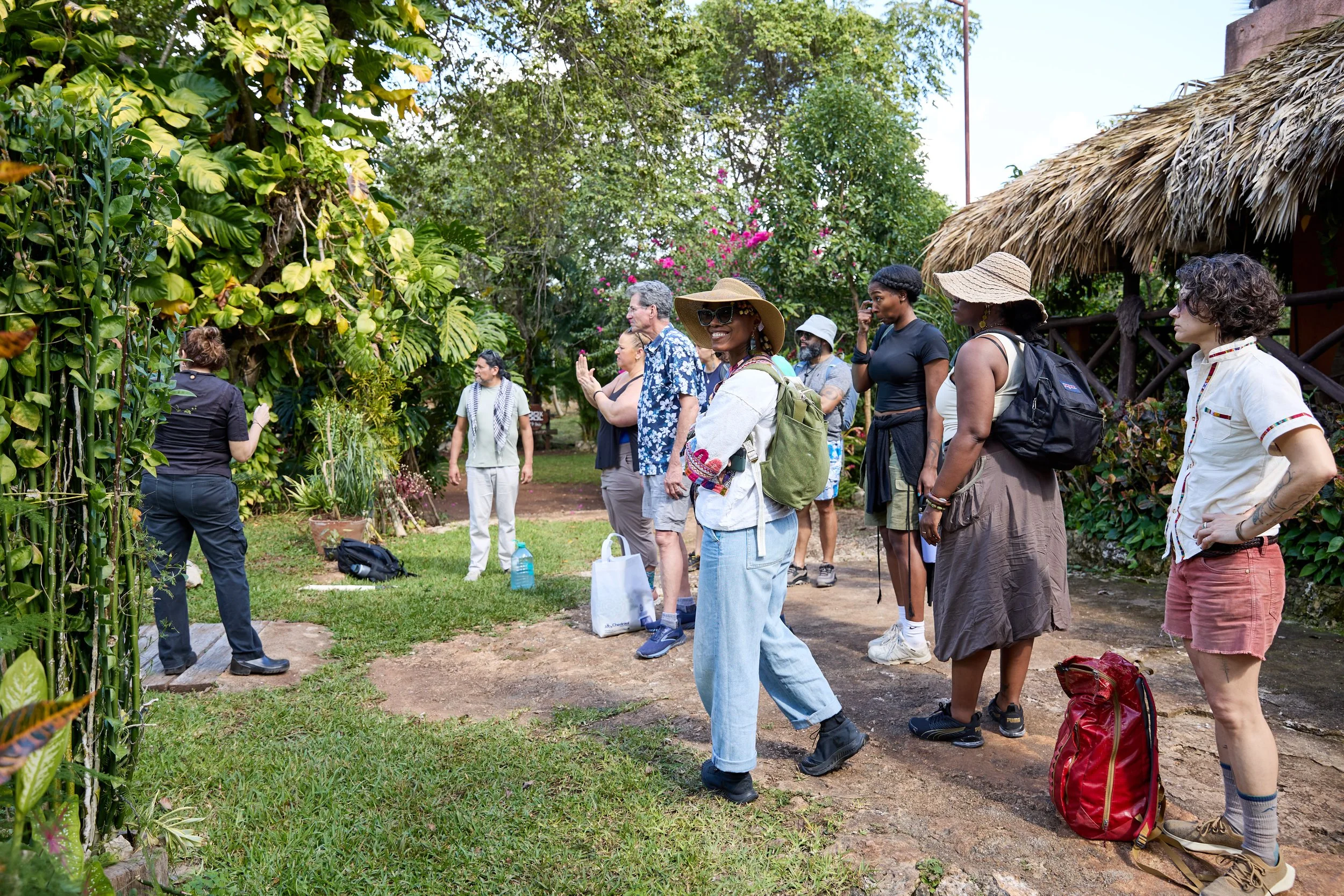 Group of people observing a garden or plant exhibit outdoors, with greenery and thatched roof structures in the background.
