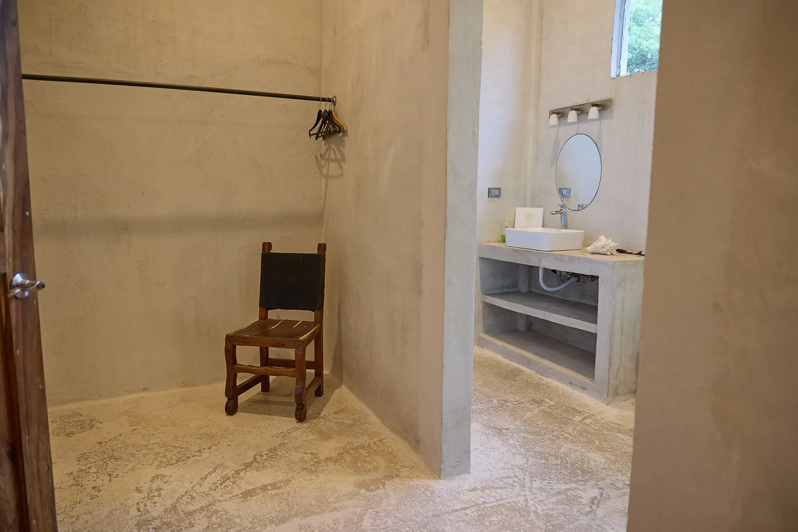 A minimalist room with a wooden chair, a clothing rod with hangers, and an adjacent bathroom area with a sink, mirror, and window.