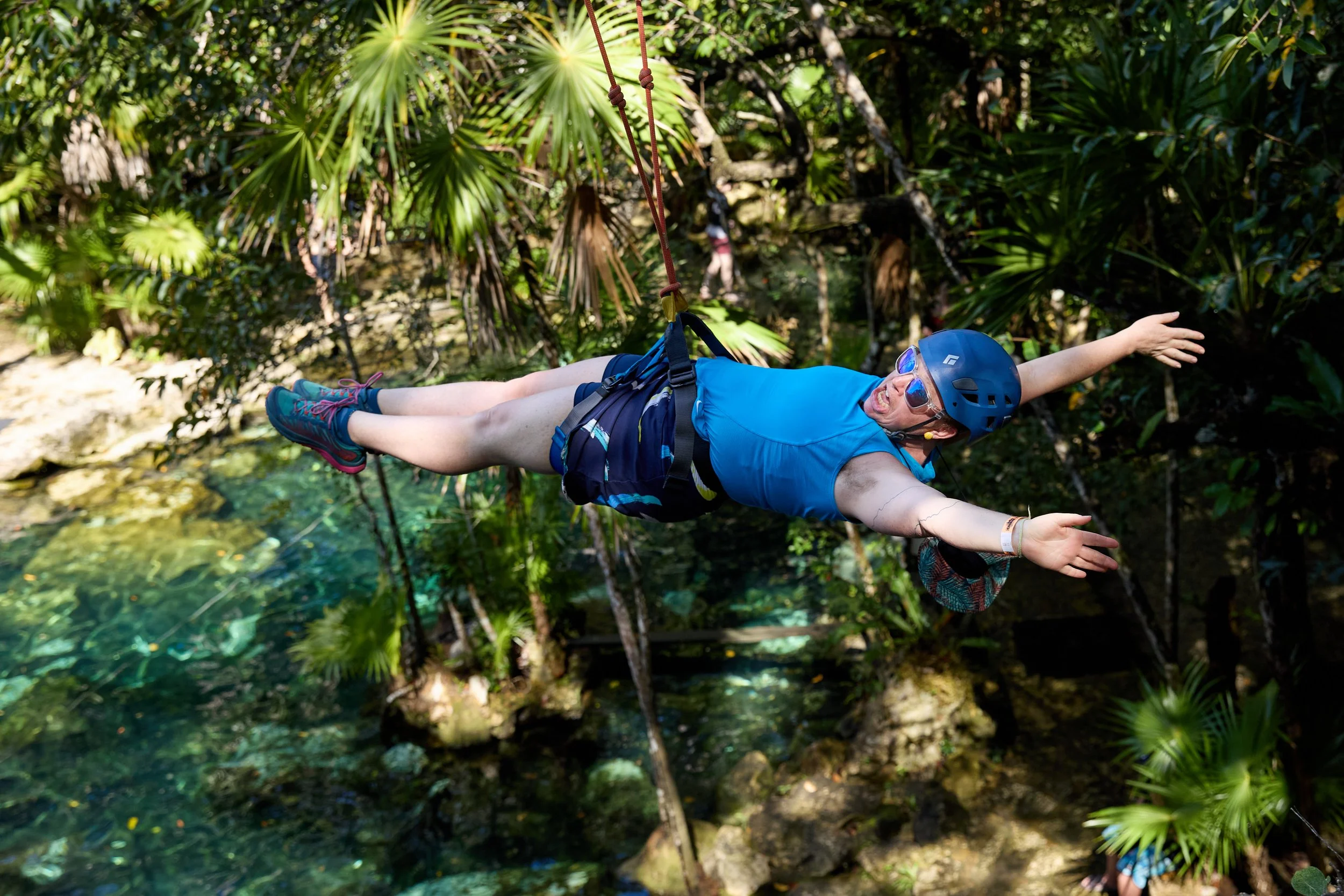 Person wearing a blue helmet and blue shirt ziplining over a lush jungle with water below.
