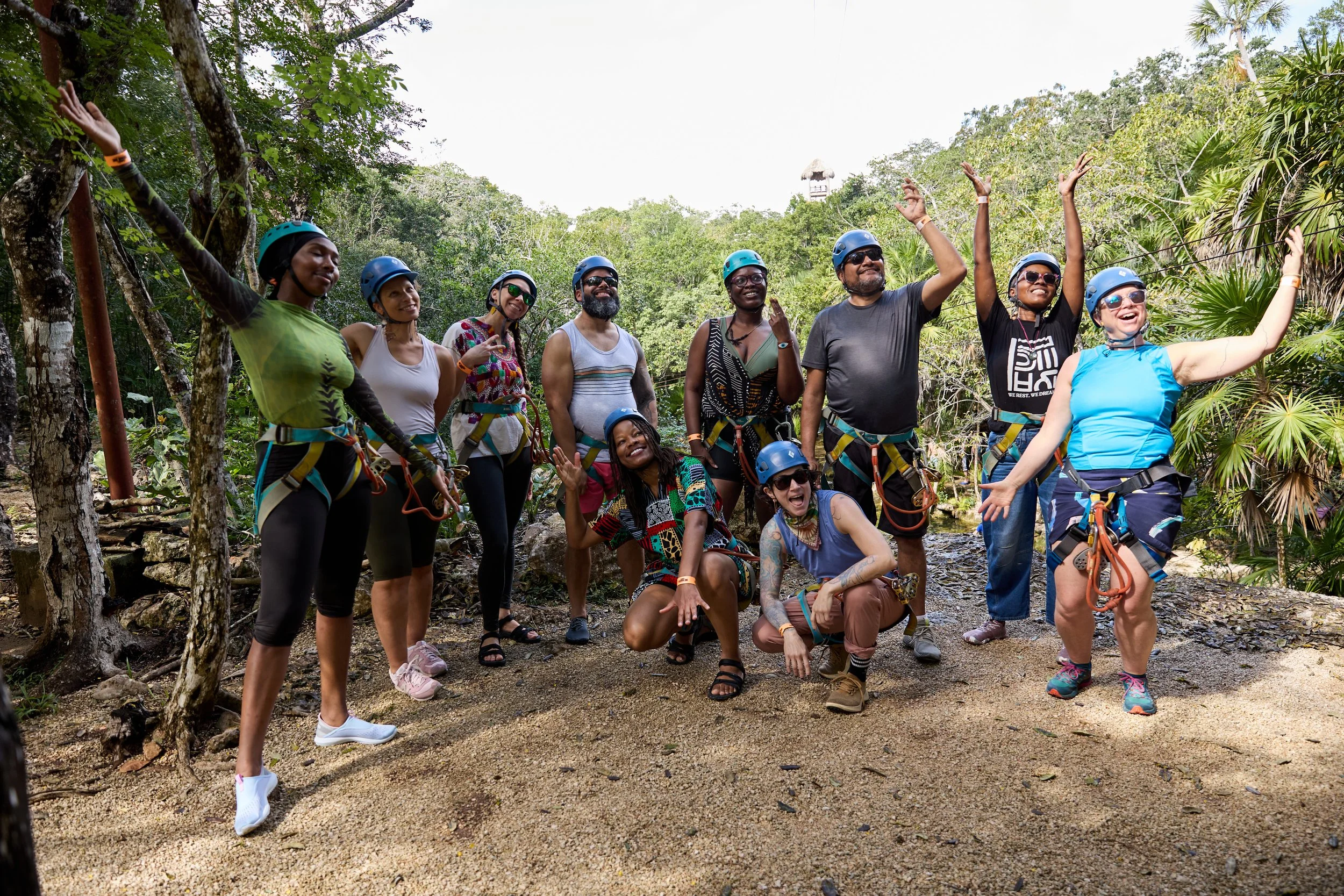 A group of people wearing helmets and harnesses for outdoor adventure, standing on a dirt path in a lush green forest, smiling and posing.