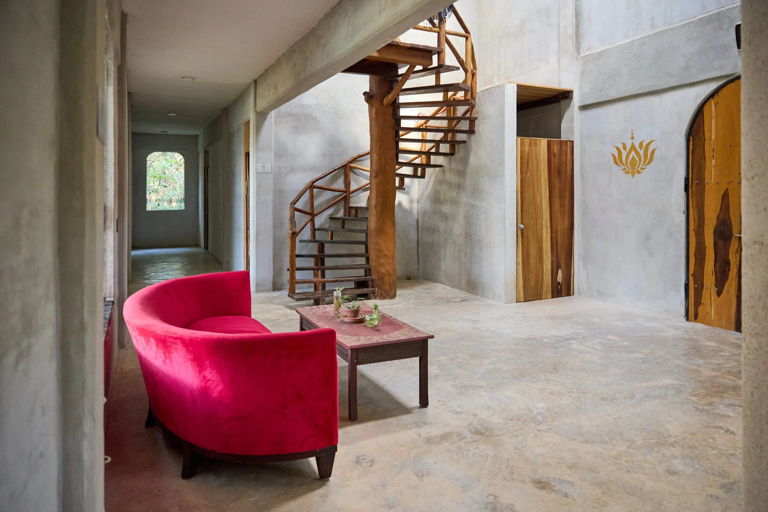 Interior of a minimalist home with concrete walls and floors. A red velvet curved sofa is positioned near a small wooden coffee table with plants and cups. There's a wooden spiral staircase leading to an upper level, and wooden doors on the walls. Na