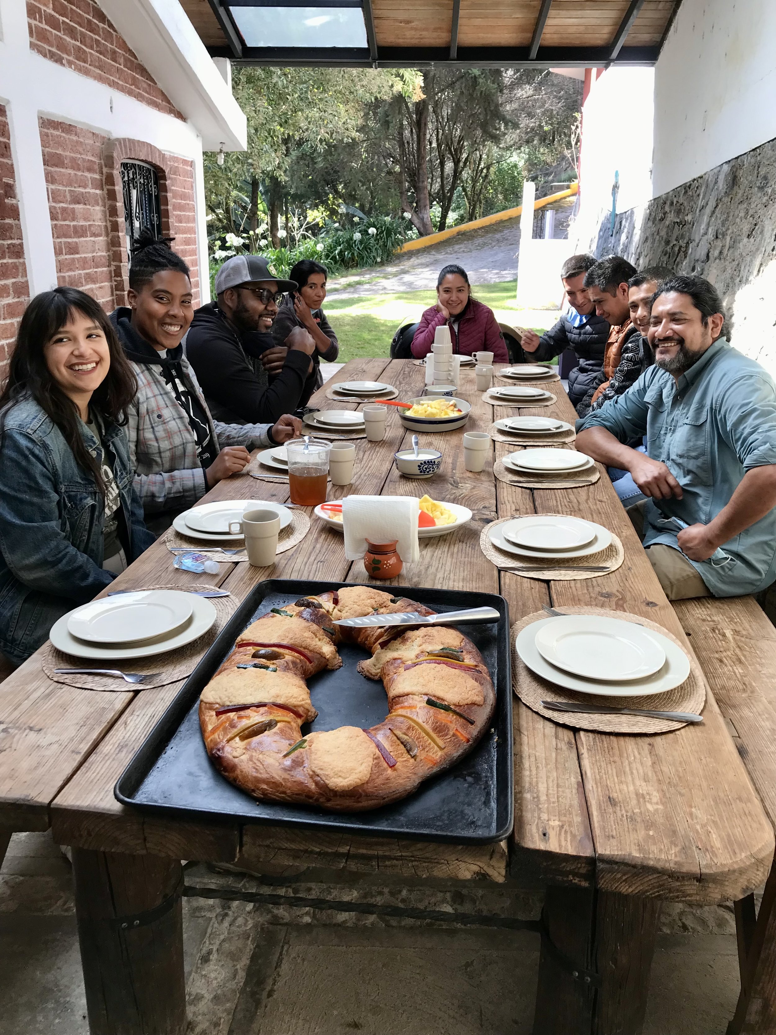 A group of nine people sitting around a wooden outdoor dining table, smiling, with a large pastry in the center. The table has plates, cups, and bowls, with a tray of baked pastry ring in the foreground.