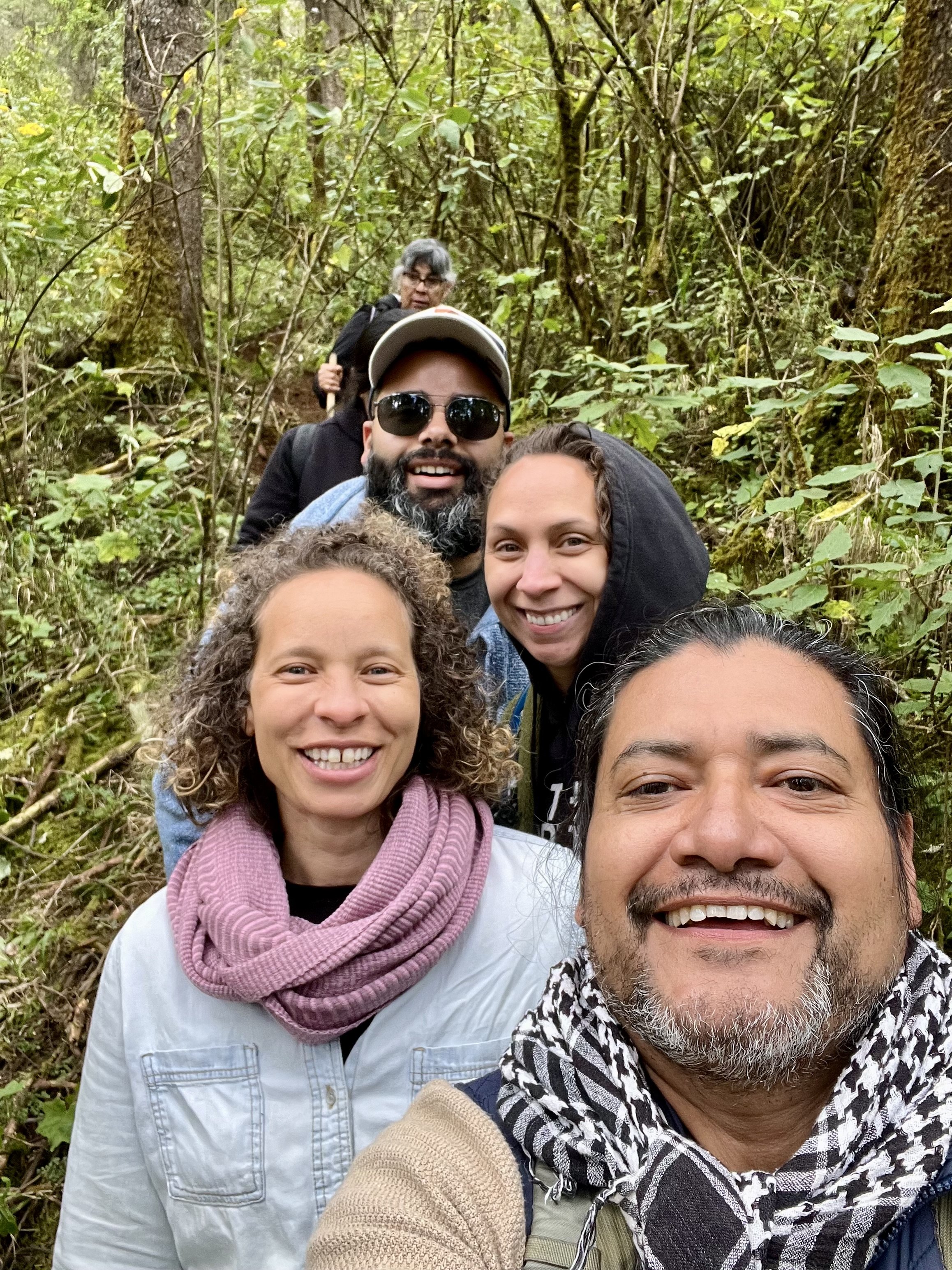 A group of five people taking a selfie on a hiking trail in a dense forest, smiling happily at the camera.