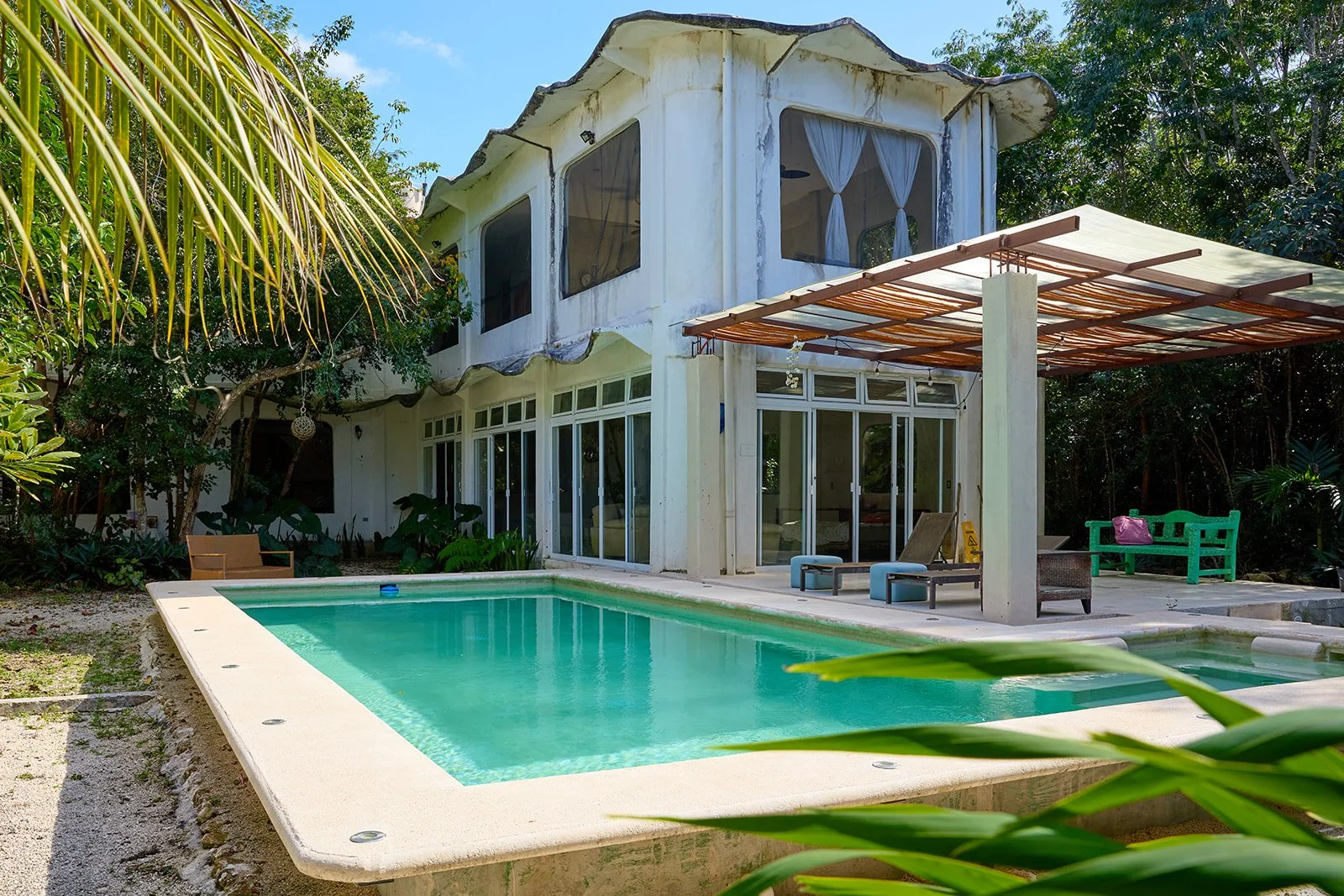 A two-story house with large windows and white curtains, surrounded by lush green trees and plants, with a rectangular swimming pool in the foreground and outdoor furniture on the patio.