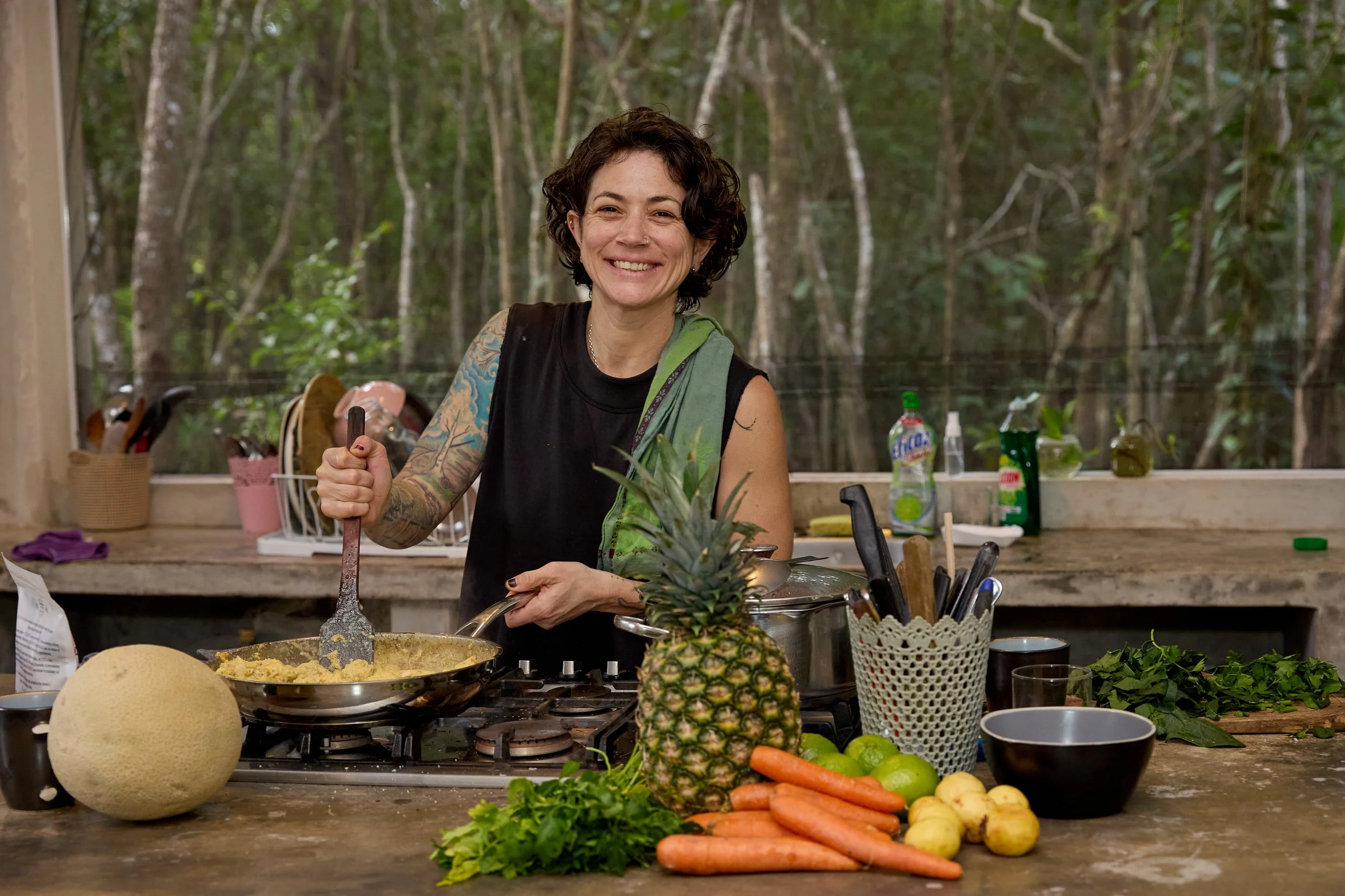 A woman with short curly dark hair and tattoos on her arm, smiling while cooking in a rustic outdoor kitchen with a wooded background. The kitchen counter has various fresh vegetables including carrots, potatoes, a pineapple, and green limes, along w