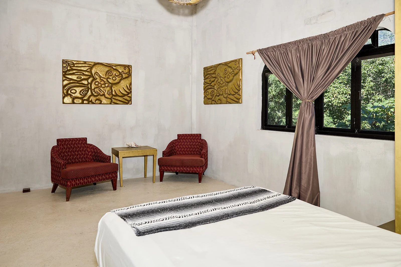 A hotel room with two red armchairs, a small wooden table, two gold decorative wall hangings, a window with brown curtains, and a bed with a black and white striped runner.