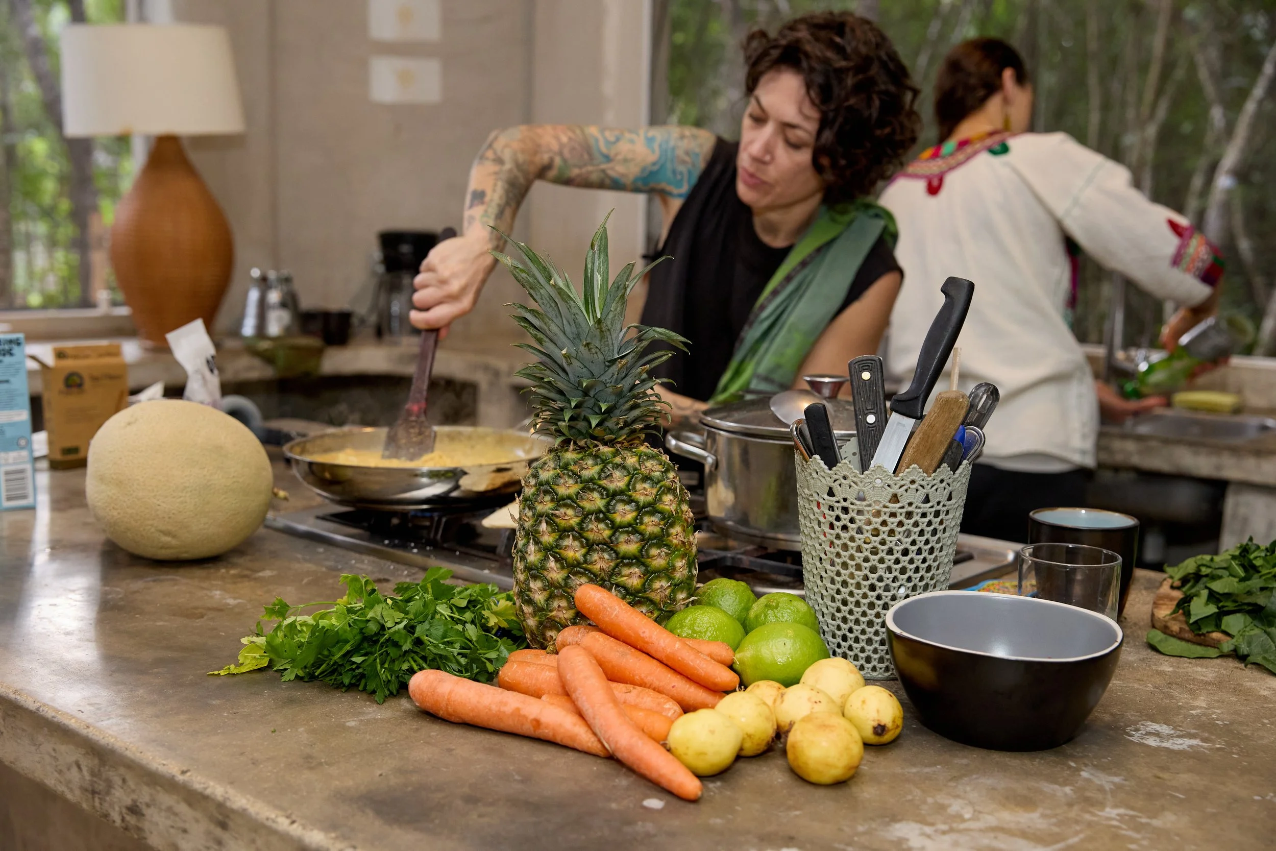 Two women preparing food in a kitchen surrounded by fresh produce including pineapple, carrots, limes, apples, and melon, with kitchen utensils and a blender visible.