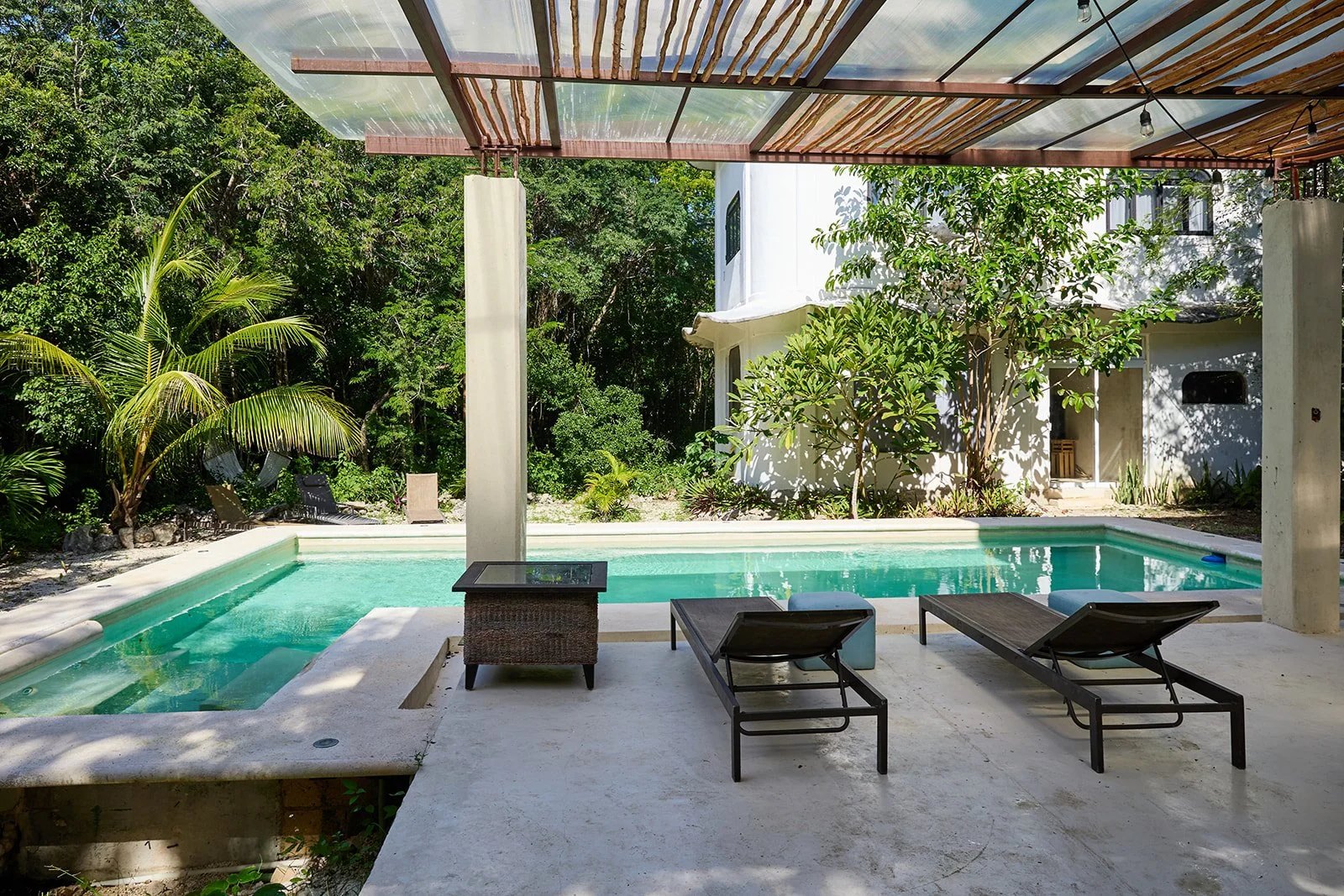 Modern poolside patio with lounge chairs, a wooden table, lush greenery, trees, and a white building in the background, shaded by a transparent roof.