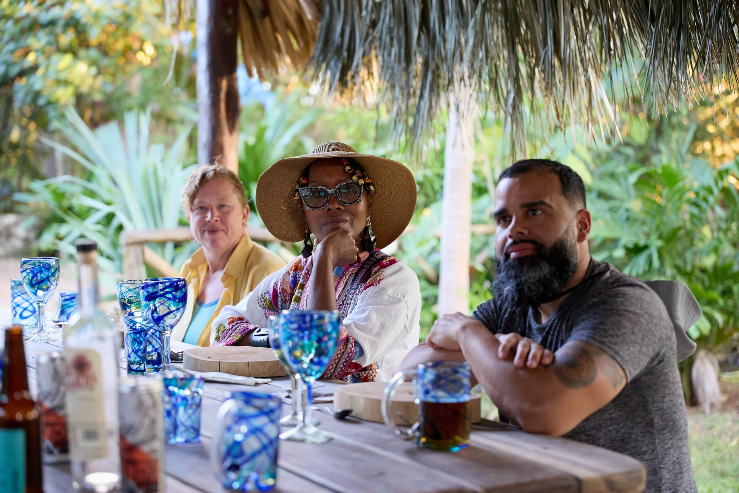 Three people sitting at a table outdoors in a lush tropical setting under a thatched roof, with glasses and bottles on the table.