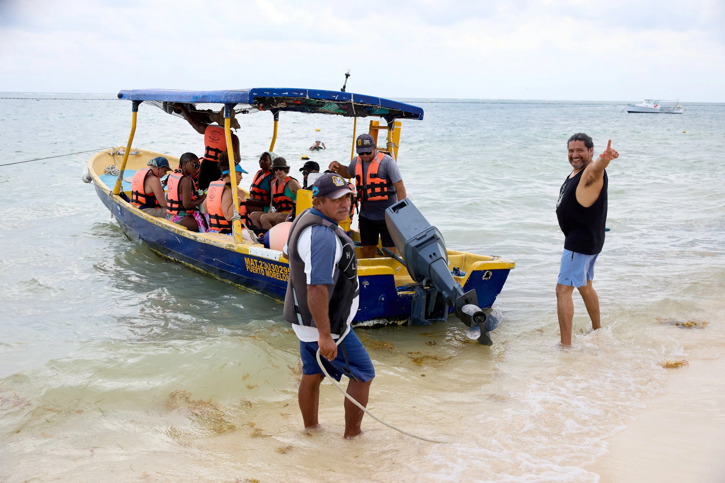 Group of people on a boat and in the water at the beach, with two men standing in shallow water.