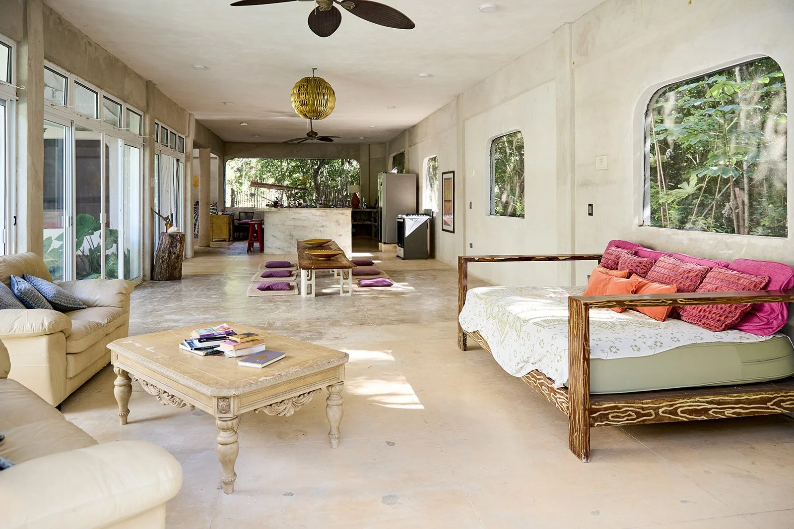 Open living room with large windows showing greenery outside. Features include a beige leather sofa with pillows, a wooden coffee table with books, a daybed with pink and orange cushions, a dining table with purple cushions, a fan hanging from the ce
