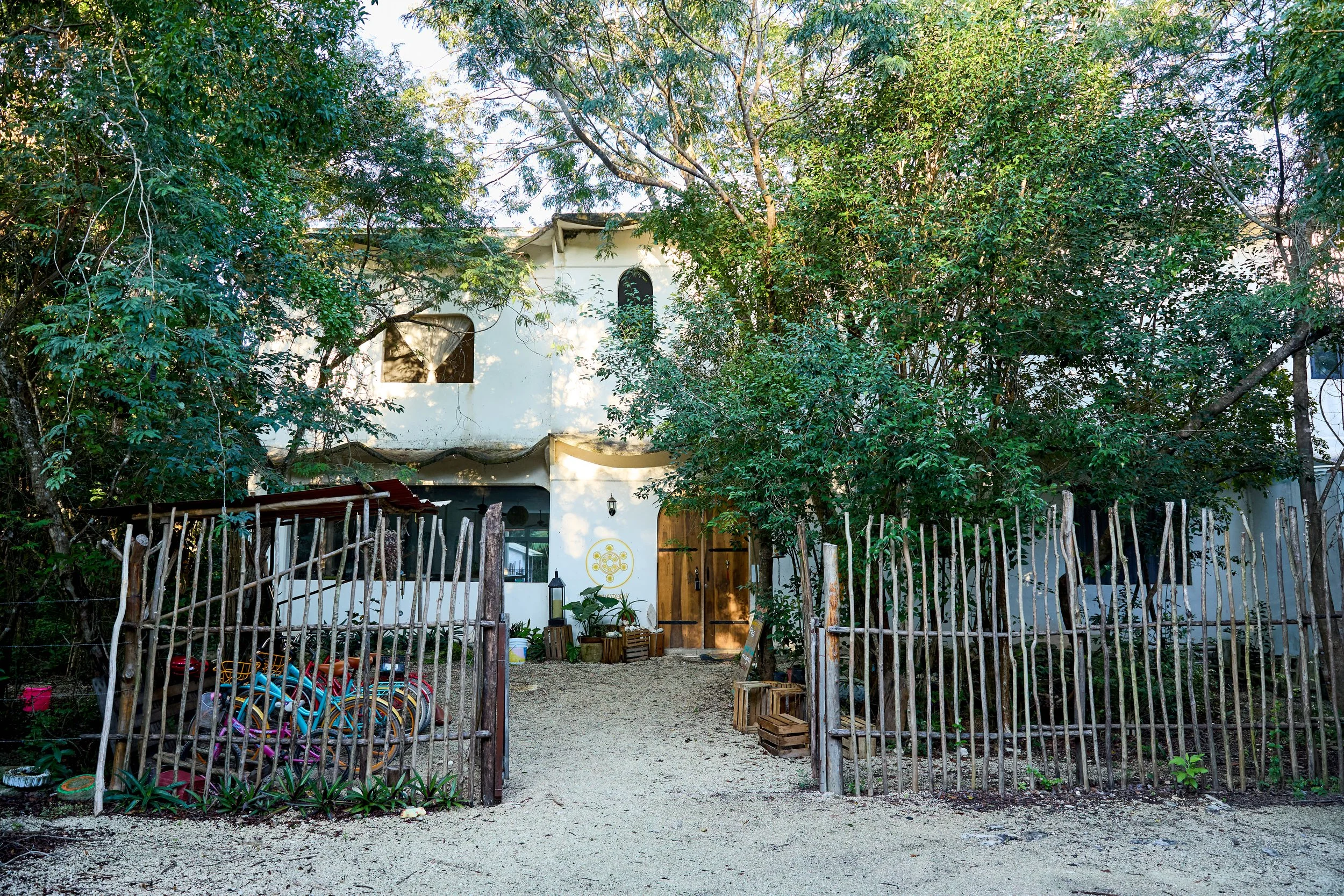 A white, two-story building with wooden door, surrounded by trees and wooden fence, with bicycles and garden decor in front
