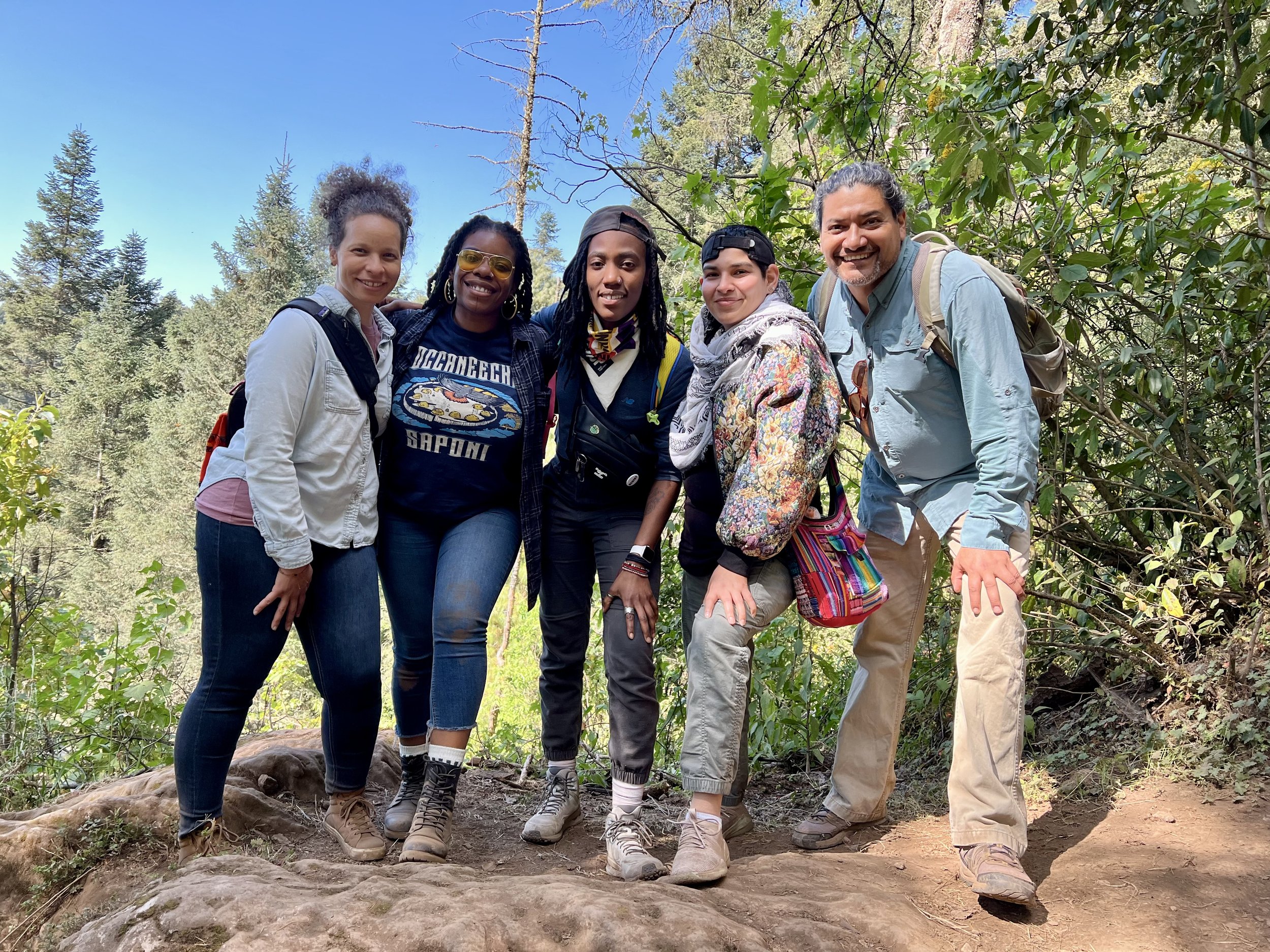 Group of five diverse people standing together outdoors in a forest, smiling for the camera, on a hiking trail surrounded by trees and greenery.