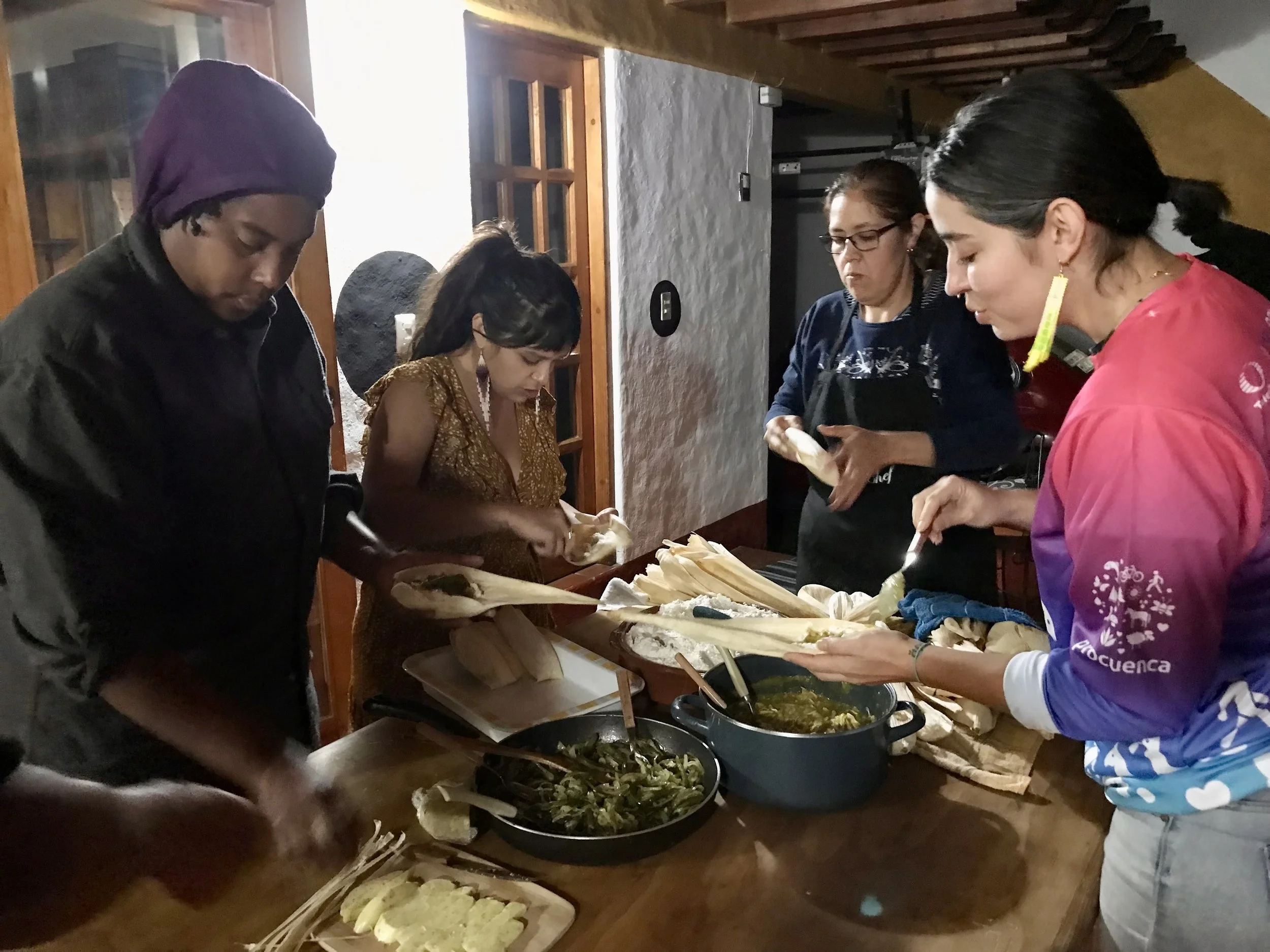 Four women preparing food together in a kitchen, wrapping tortillas with filling.
