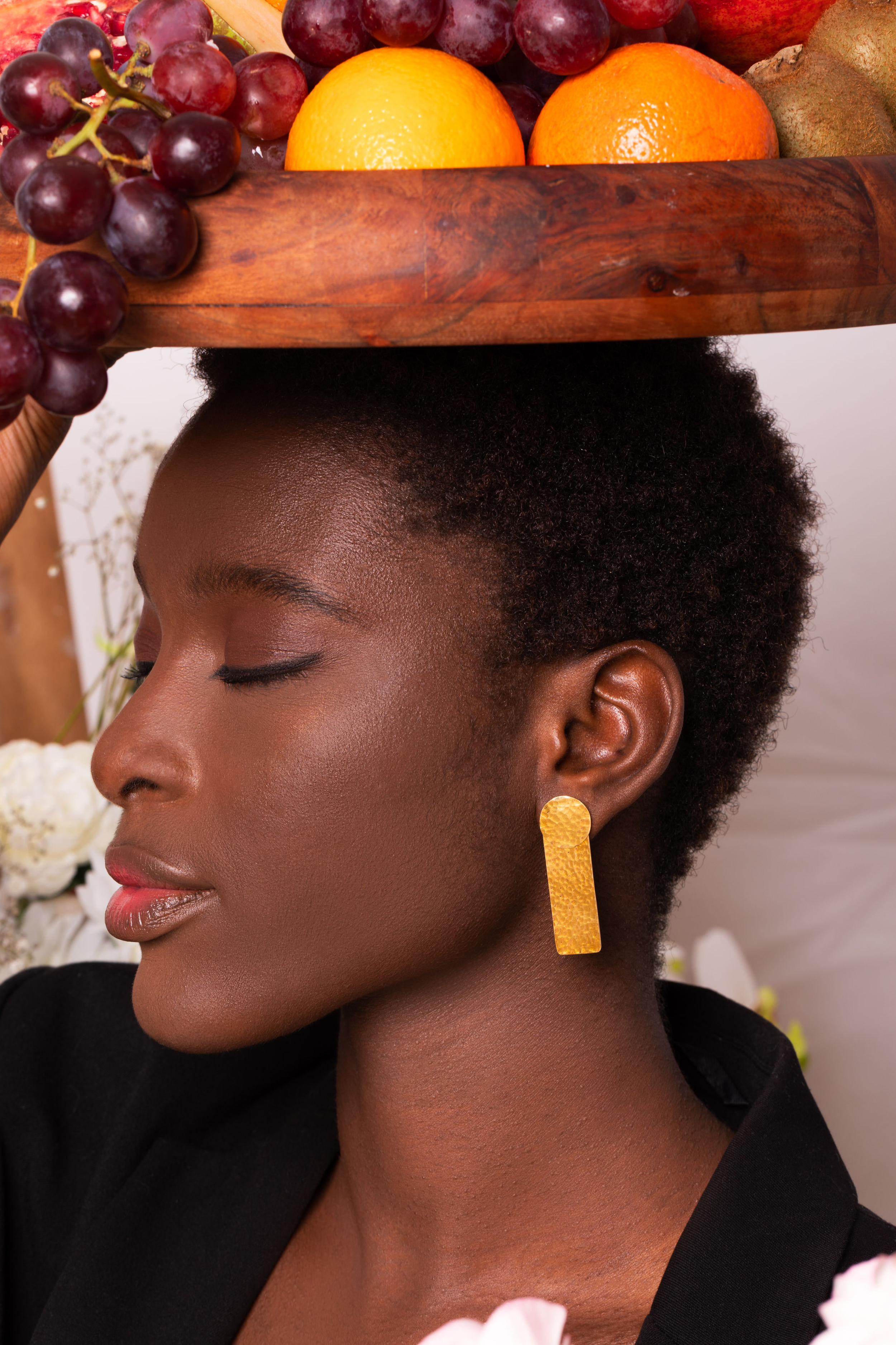 A woman with dark skin and short curly hair wearing gold earrings and a black outfit, balancing a wooden tray with oranges, grapes, and other fruits on her head, with closed eyes.
