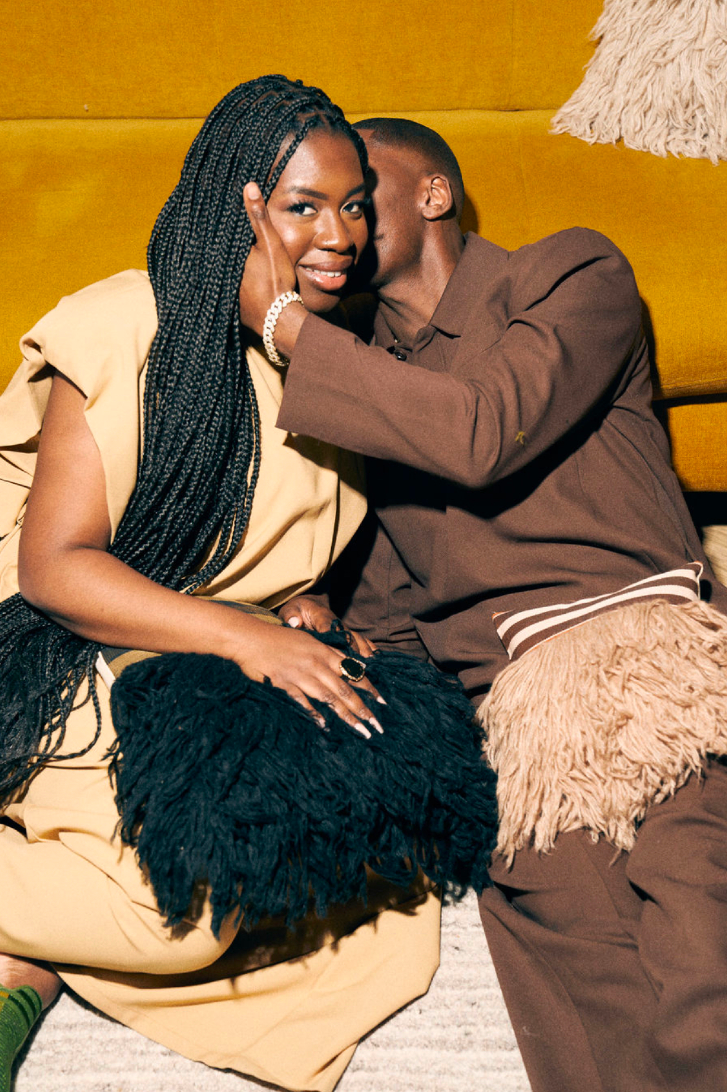 A man whispering into a woman's ear on a mustard-colored sofa, with the woman smiling and looking at the camera, in a cozy indoor setting.